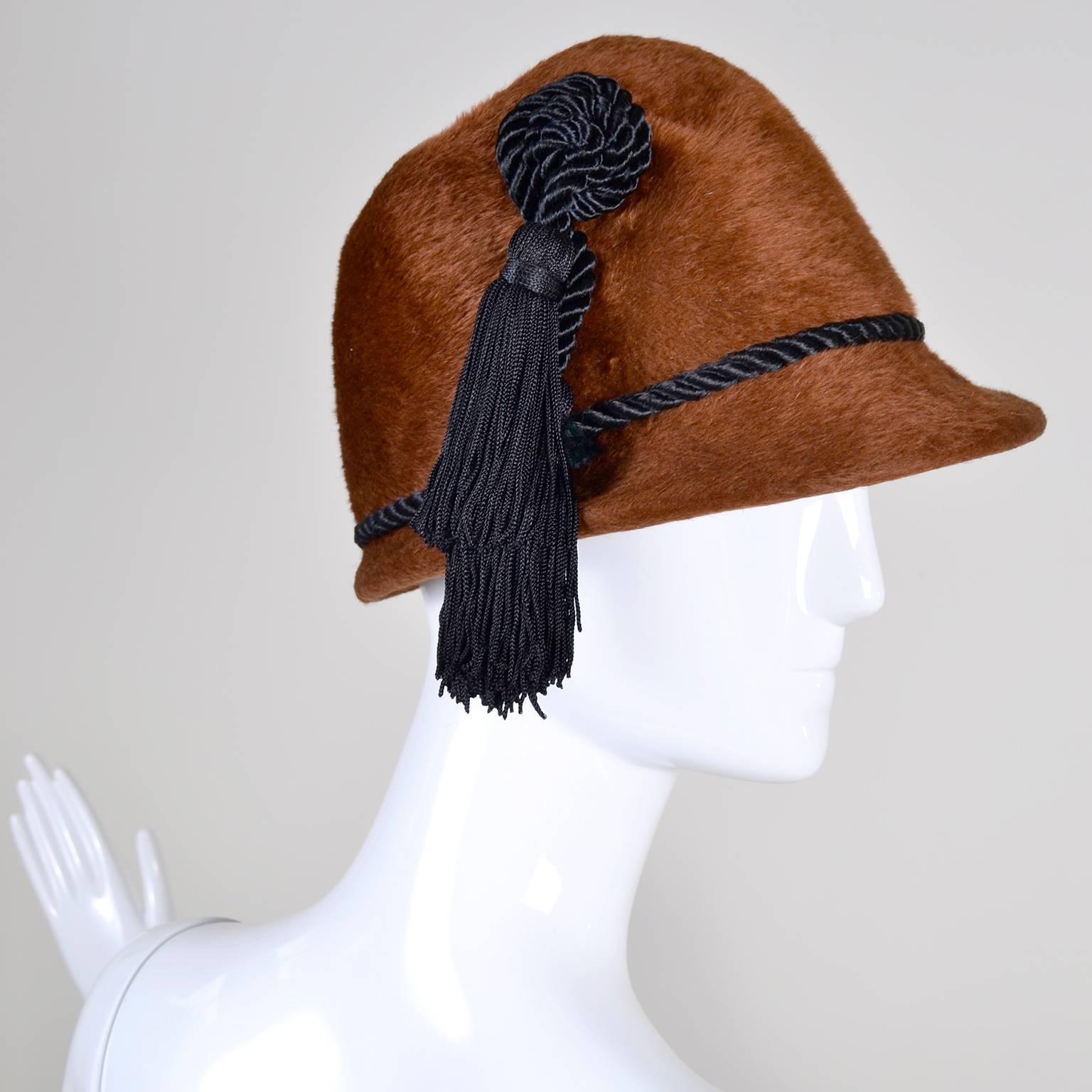 This is a luxurious brown faux fur vintage YSL hat with a short brim in front and a black braided cords There is a thin cord around the hat, and another black cord ending in two large black tassels that hang down past the brim. This hat is cloche