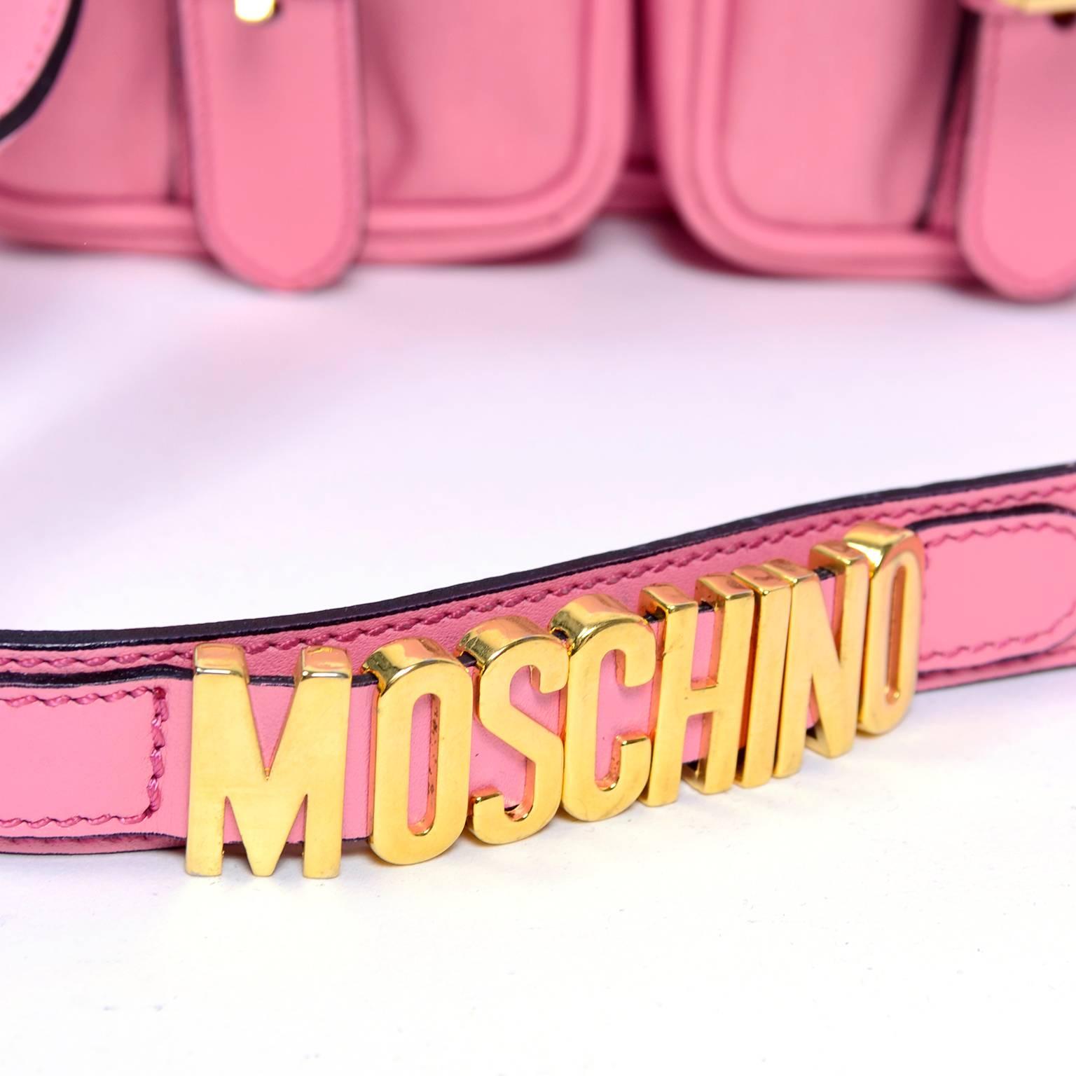 This is an iconic pink Moschino handbag with gold hardware. The top flap comes over to snap over the two front pockets, with faux buckles on the front, for a school satchel look. This nylon bag is lined with black logo fabric. The strap is