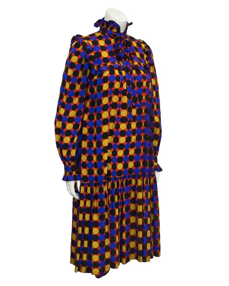 1970's YSL full cut dress with black, royal blue and yellow polka dot pattern on red background. Full swing fit with drop waist and gathered hemline. Ruffled trim on the cuffs. Tie at neck and gathered yoke with inset ruffle detail extends to the