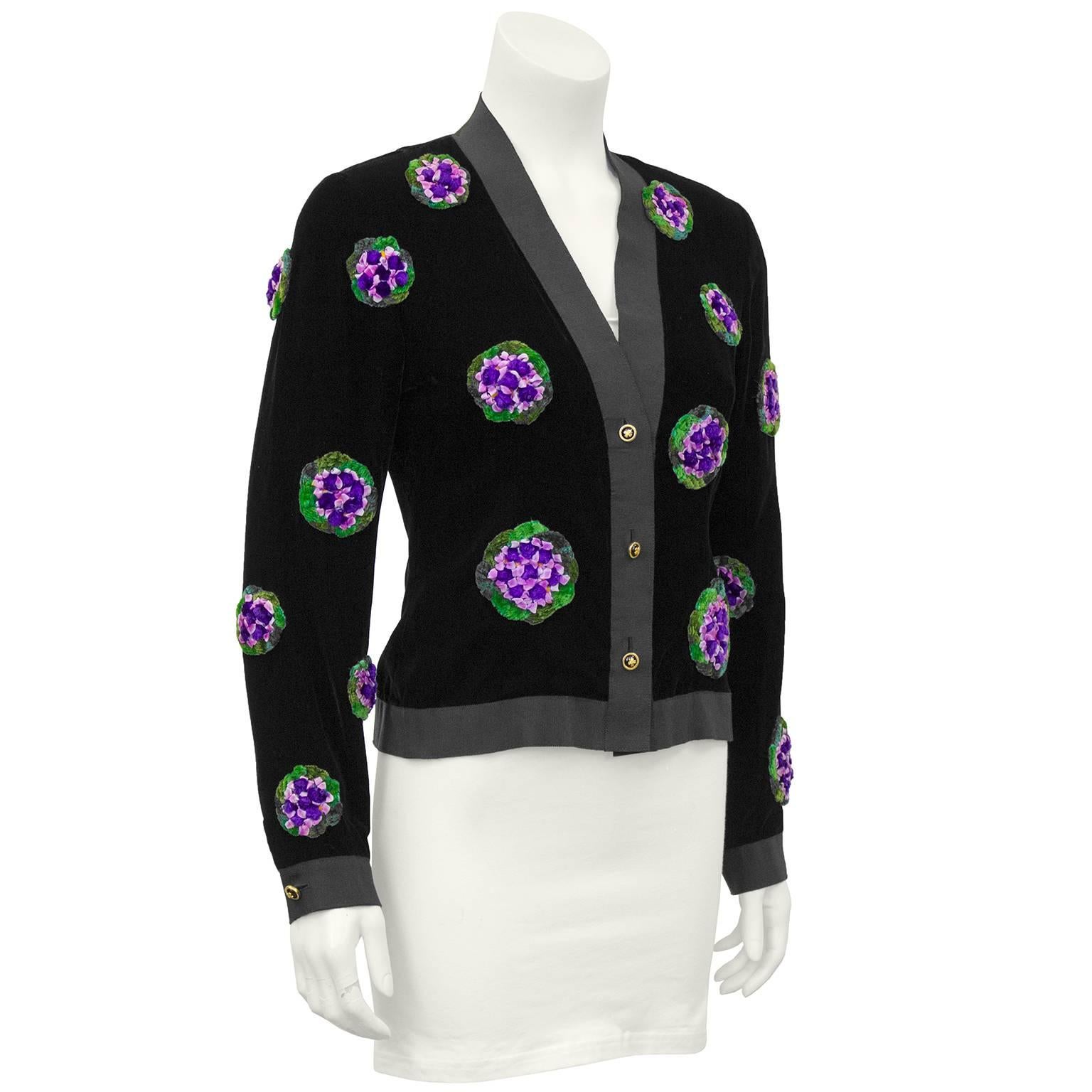 1990s Chanel jet black velvet cardigan featuring black grosgrain ribbon trim, 3D purple and green floral appliques and gold Chanel logo buttons. Great seasonally transitional piece. Excellent vintage condition, fits like a US 6. 

Sleeve 21"