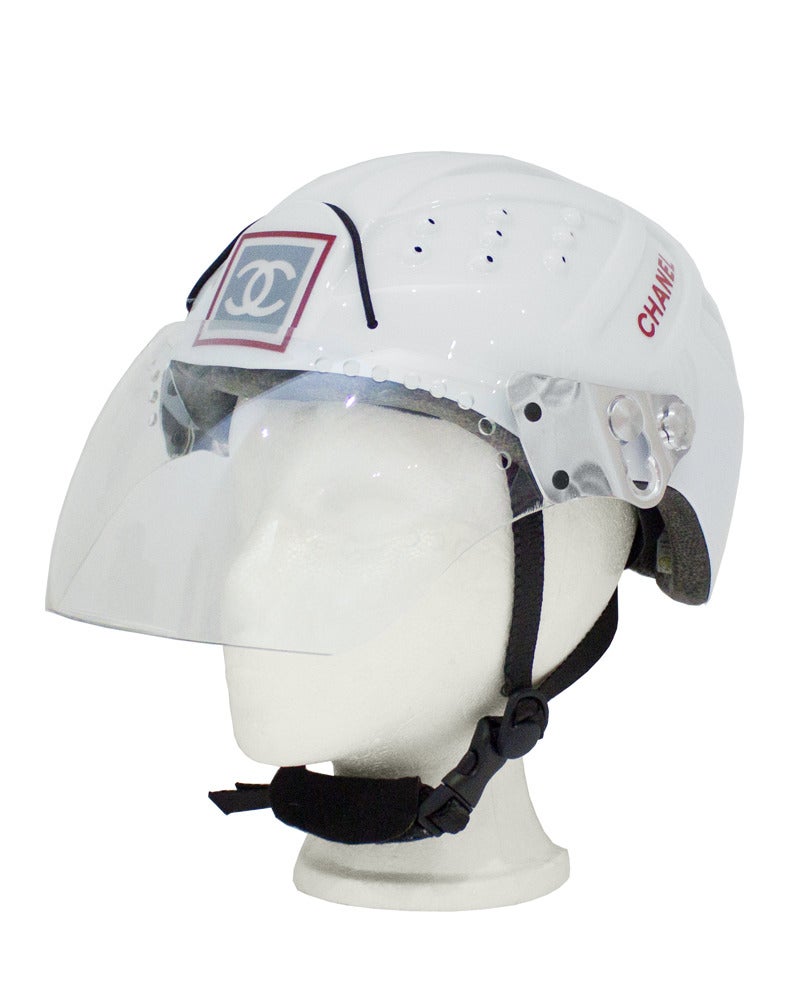 1990's white Chanel helmet that can be used (as stated by sticker on inside of helmet) for mountaineering and ice climbing. Plastic shell with styrofoam interior. Adjustable black buckle strap closure at chin. Clear visor covers face with two