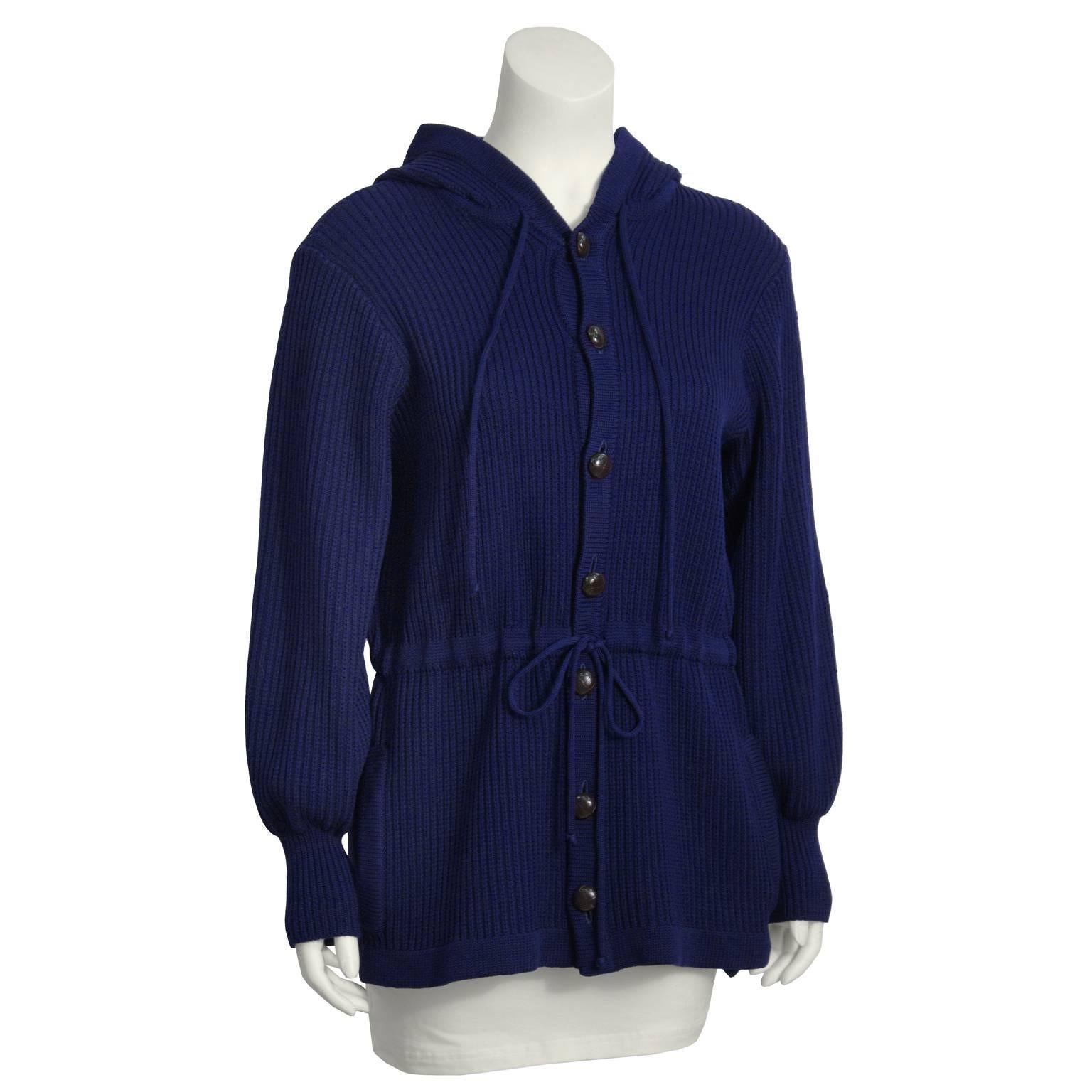 Stay cozy and chic with this Yves Saint Laurent navy hooded knit cardigan from the 1970s. Features drawstring at neck and waist. Two slit pockets at the hips. Front button closure. Brown buttons have woven motif. Sleeves are loose fitting with