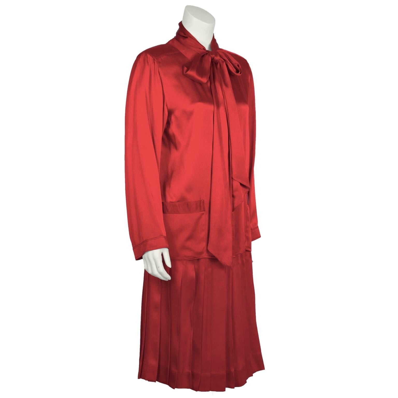 1980's Chanel red satin ensemble featuring a blouse with self tie bow and a stitch pleated. The blouse features a banded cardigan-style neckline with gold CC buttons down the front padded shoulders, and front hip pockets. Pleated fitted skirt
