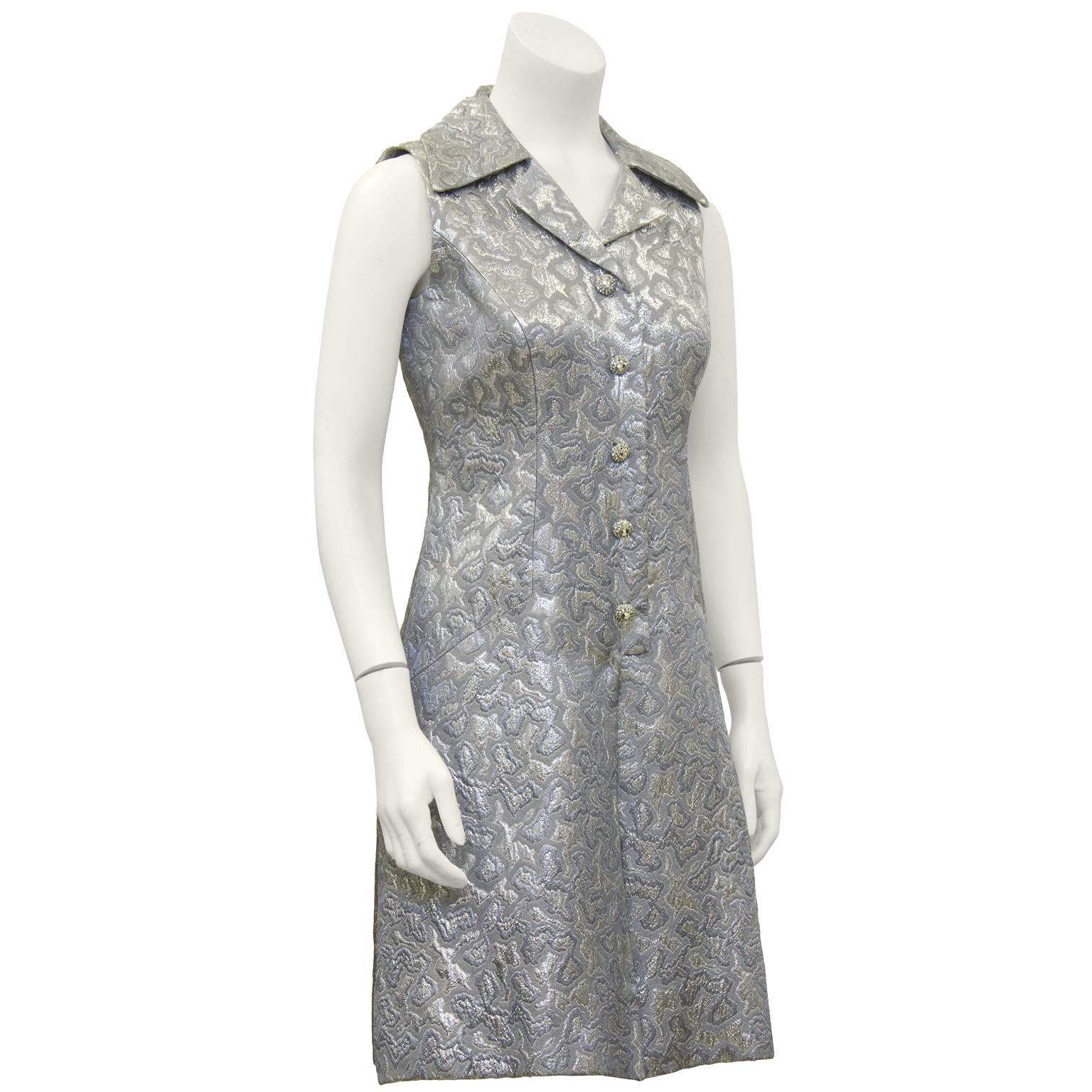 1960's Adele Simpson sophisticated and sporty silver brocade sleeveless dress. Dress has an oversized notched lapel and rhinestone encrusted buttons. Drop style waist with inverted front pleat. The brocade fabric features a silver blend with an