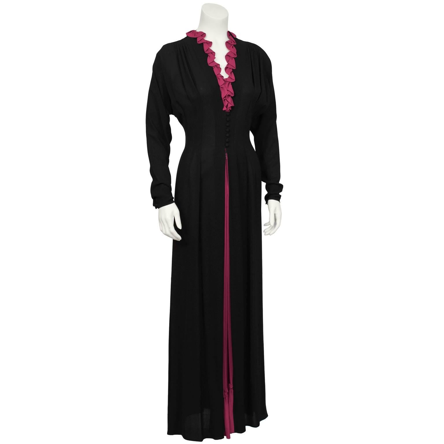 Black moss crepe tea gown with fuchsia ruffle detail from the 1970’s. Dress features dolman style sleeve and a deep V neckline detailed with a bold fuchsia ruffle. Fitted at the waist, fastens up the front with 7 covered buttons and a hidden zipper.