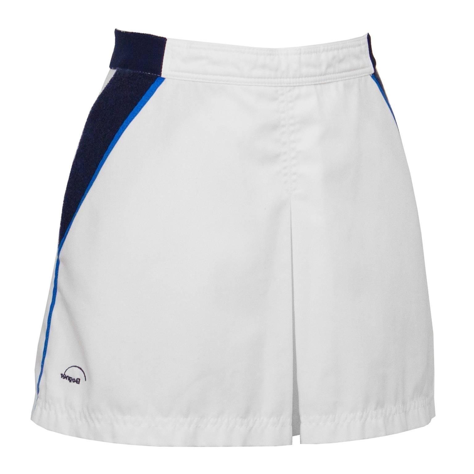 White cotton Bogner tennis skirt from the 1970's. White and navy banded waist, navy terry cloth side pockets and royal blue piping down the sides. One inverted front pleat gives the illusion that the skirt is shorts, finished with a navy embroidered