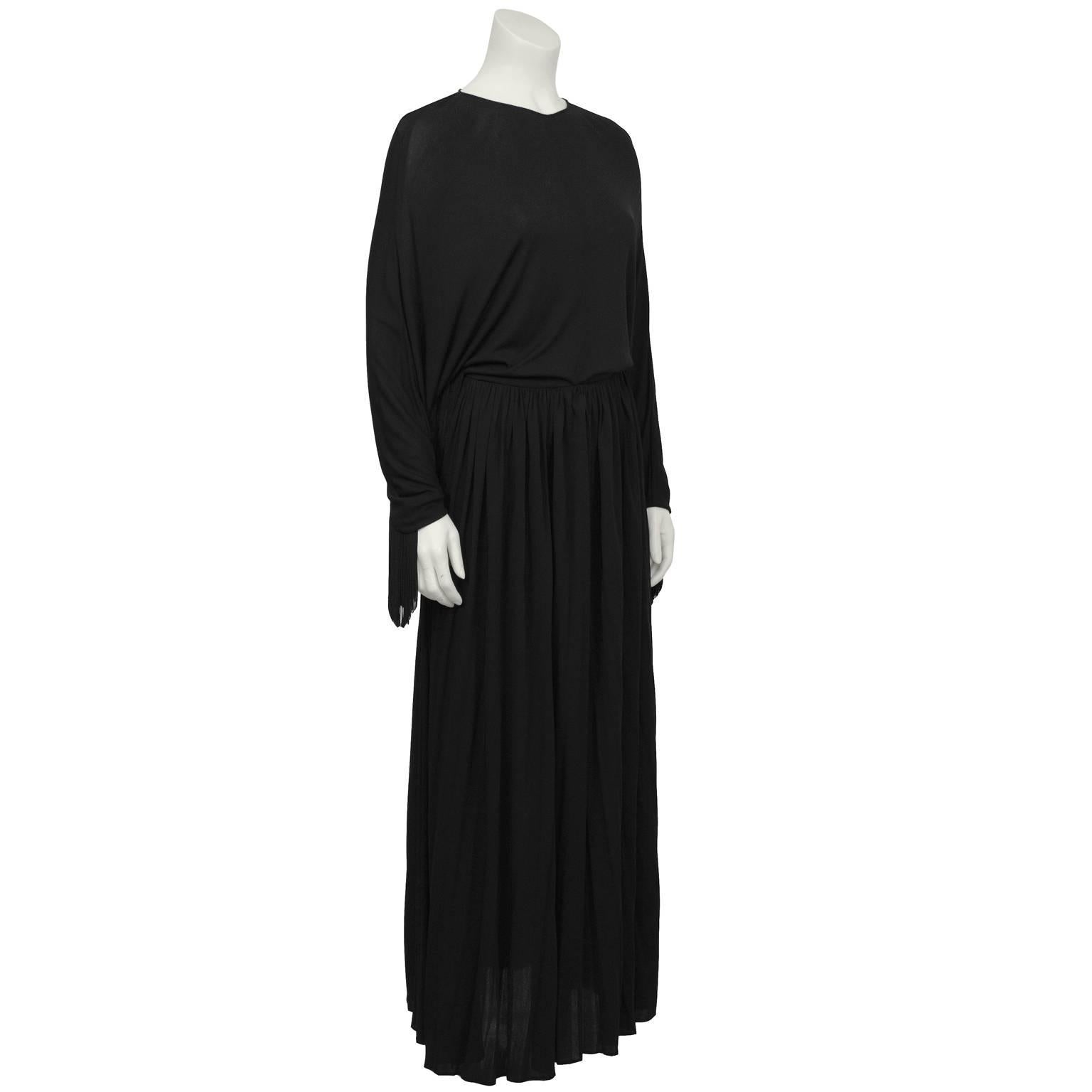Dramatic black jersey long sleeve gown from the 1970's. The gown has bat wing sleeves that are trimmed in black fringe and the skirt falls full gathered from the waistline. Zips up the back, hidden in-seam pockets and a waist tie on the inside keeps
