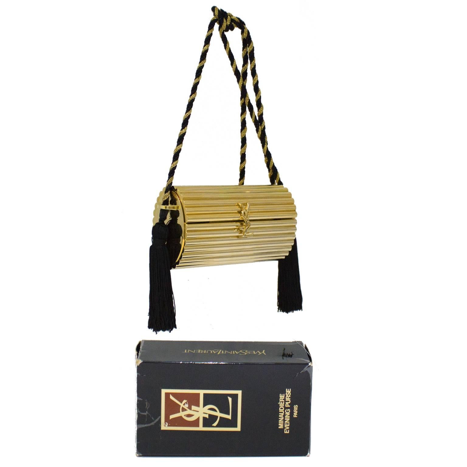 Gold metal box clutch with twisted silk cord shoulder strap by Yves Saint Laurent. The hardshell body is ribbed and shaped like a oval cylinder. Opens at the front with a hinge, YSL clasp, and is lined in gold leather. Gold and black twisted cord