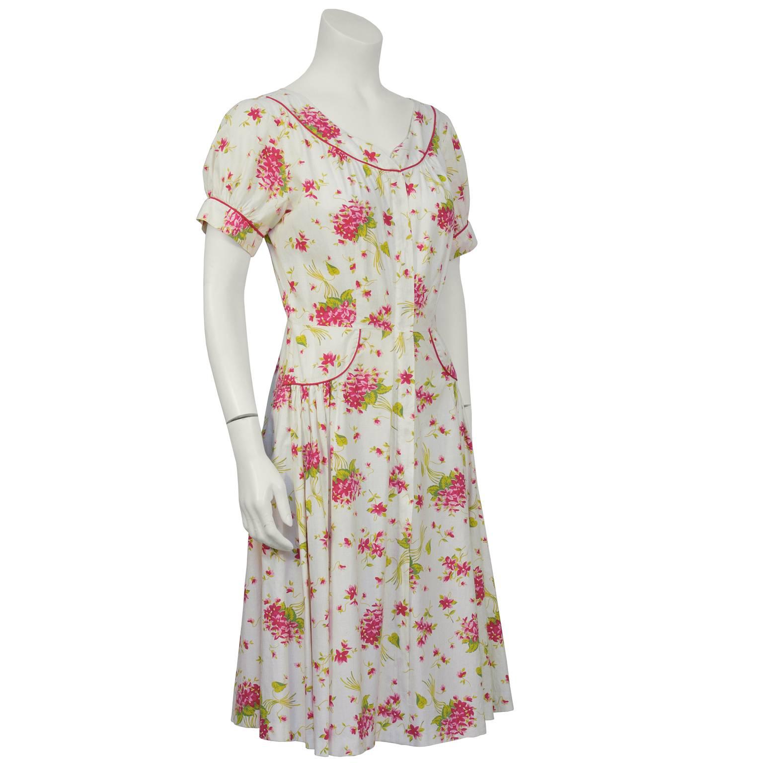 Abito da giorno in cotone con stampa floreale verde, rosa e rossa degli anni '50. Dettagli in piping rosso sullo scollo a barchetta, sui polsini e sui fianchi a semicerchio. La cerniera sul davanti è nascosta dietro un'abbottonatura. Gonna piena con