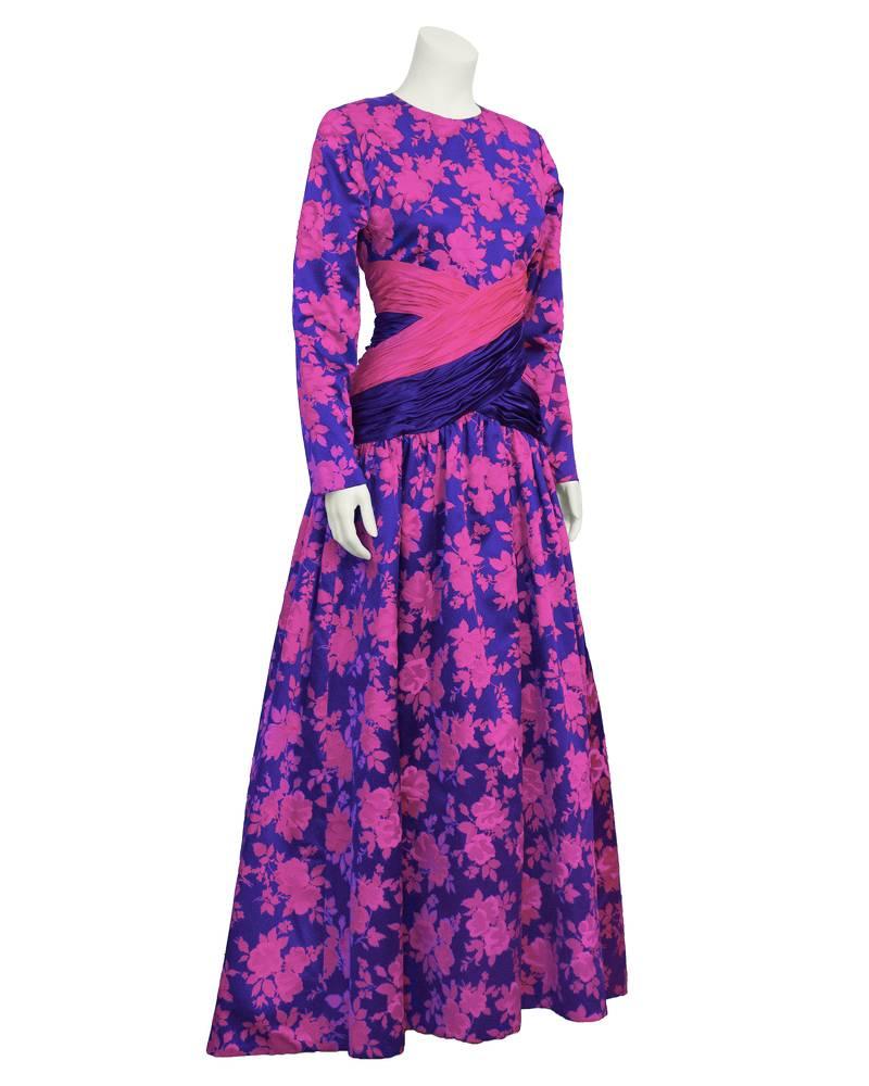 Eye-catching and vibrant 1980's gown by German designer Peter Keppler. Long sleeve, hot pink floral print on a purple background. Pink flowers are given dimension with matching pink velvet accents. Pink and purple cross over ruching at waist and