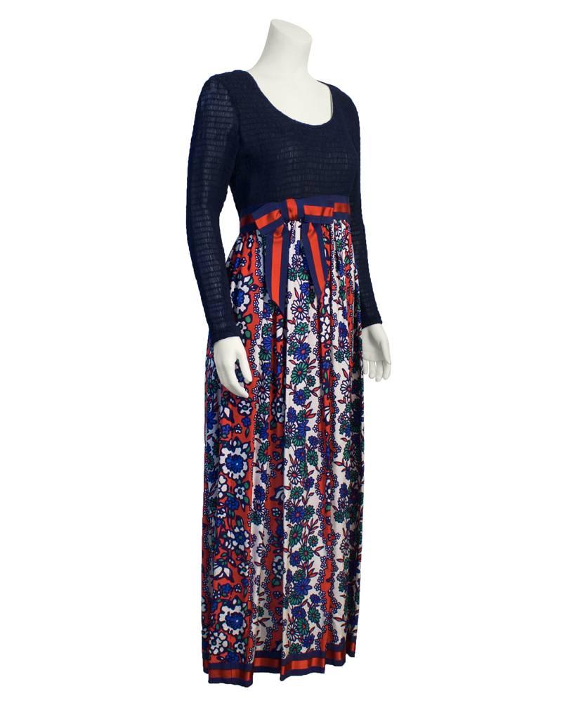 robe longue des années 1970 avec un corsage à manches longues froncé bleu marine et une longue jupe longue à fleurs rouges et bleues légèrement froncée. Taille empire avec une ceinture en ruban gros grain rouge et marine et un nœud. Une robe