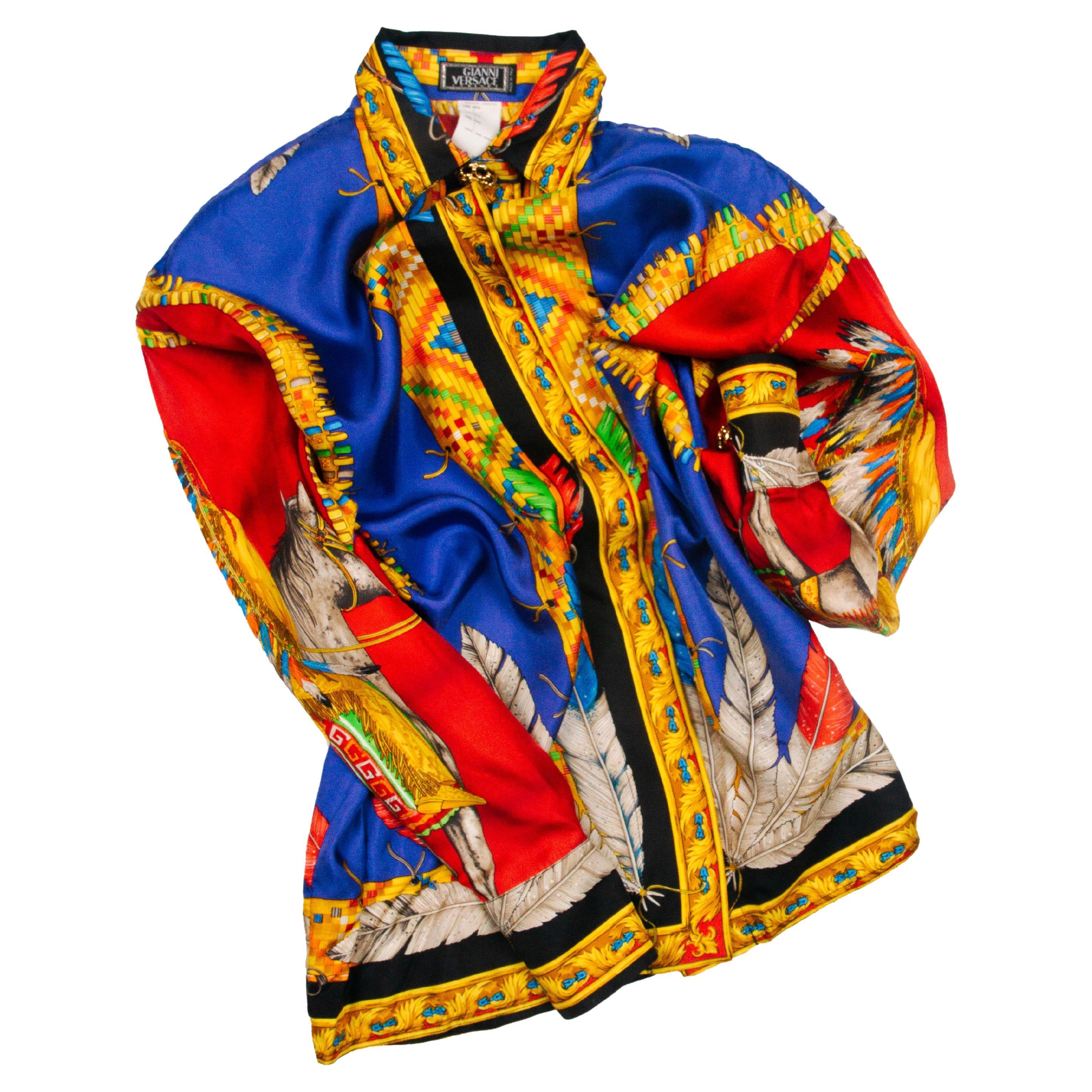 Creata da Gianni Versace per la collezione A.M.F. 1992, questa camicia di seta luminosa presenta motivi ispirati ai nativi americani, piume e una grande immagine di un uomo tribale nativo americano a cavallo. Con i suoi colori blu, rosso e giallo,