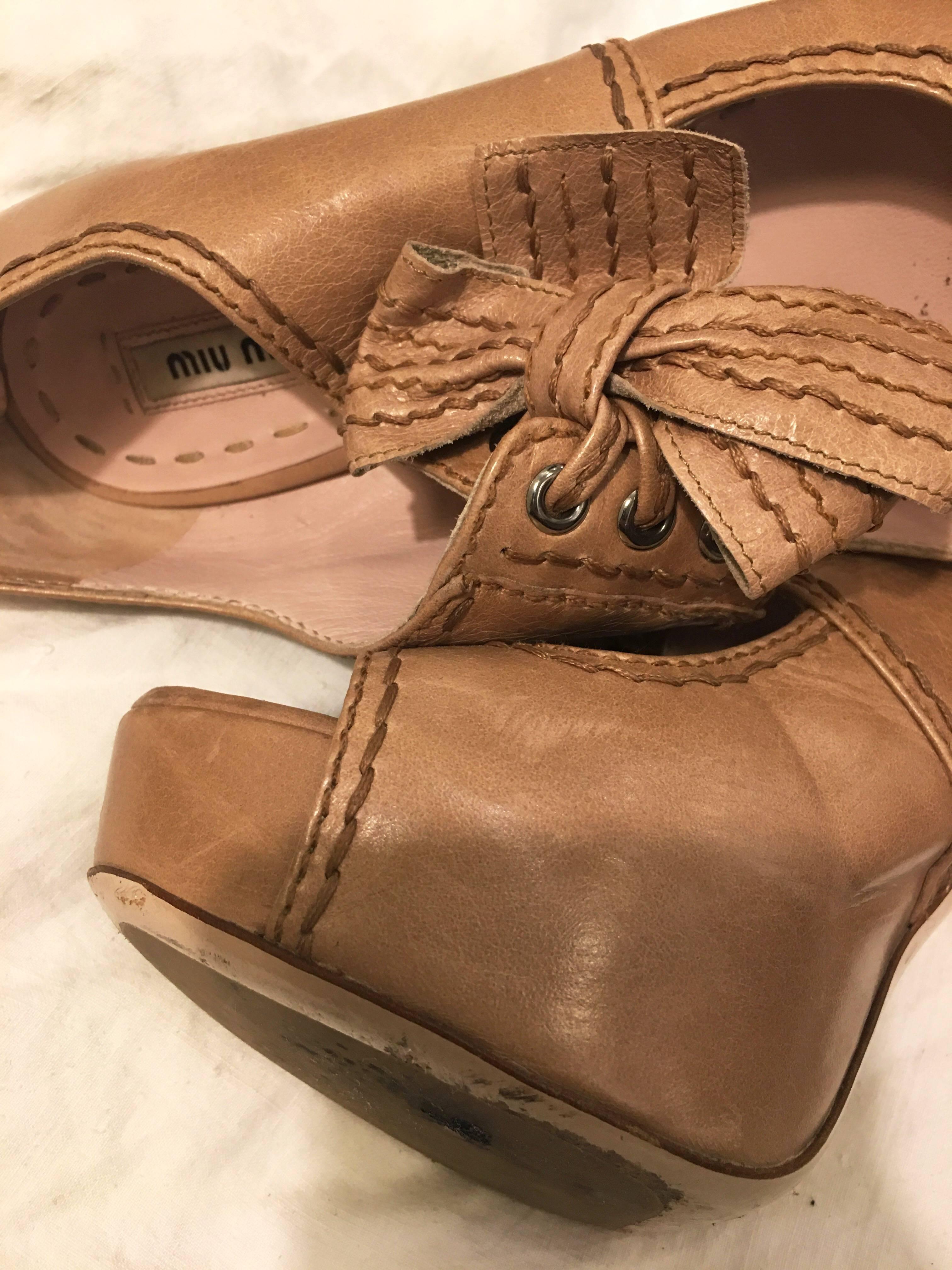 Beige Miu Miu open toe Mary Jane style pumps. Wooden stacked heels with leather laces up the front. Exposed brown stitching throughout. Zip up the back. Stamped on the bottom on the leather sole with the Miu Miu logo. 2 inch platform at toe of the