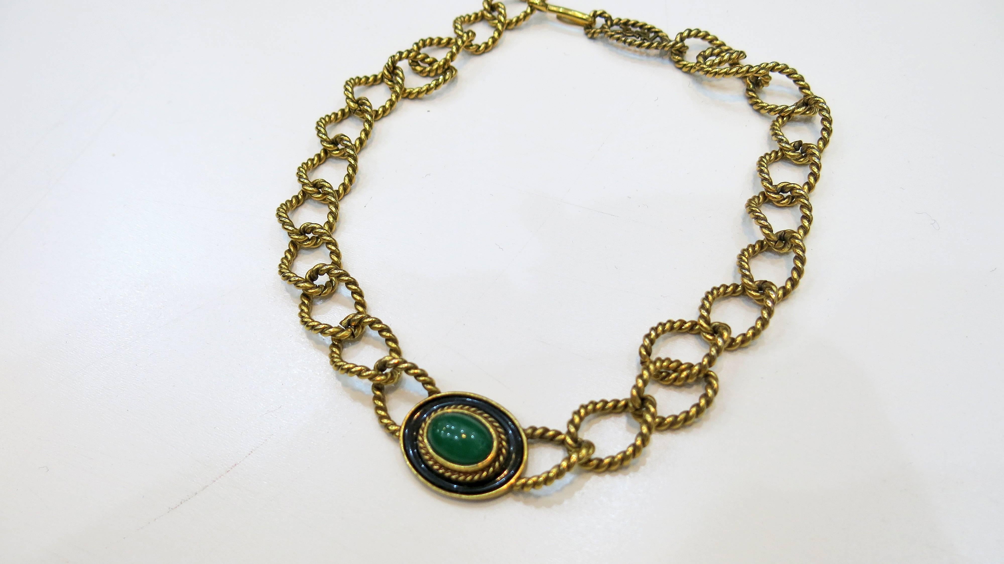 Braided brass chain with logo clasp and green glass cabochon inlaid in brass and surrounded by black enamel. Not quite a choker but hits close to the neck. 

Yves St. Laurent began as a designer making clothing for his mother and sisters. He won