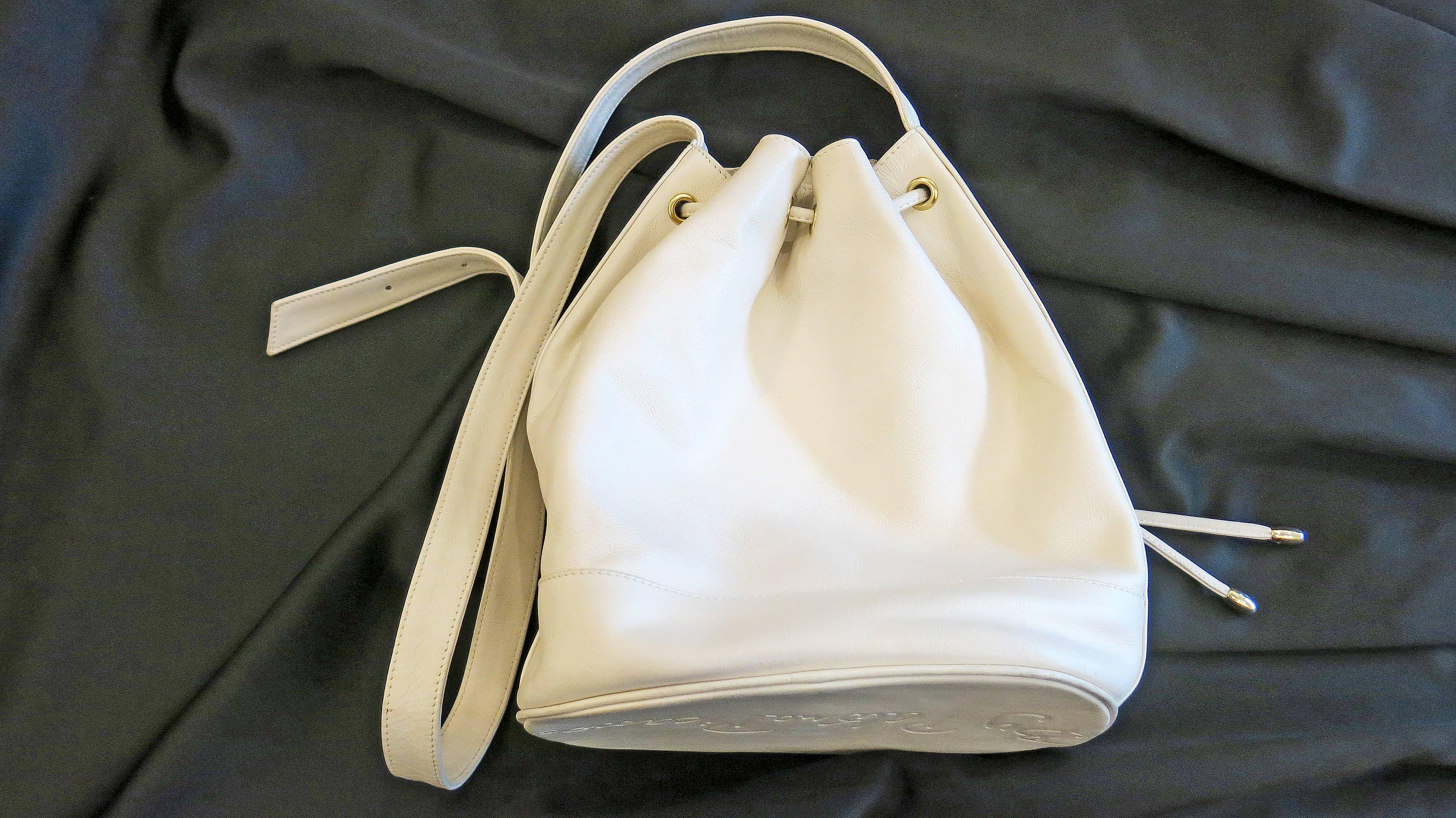White leather bucket bag with adjustable shoulder strap. Gold tone hardware. Three Paloma Picasso X logos along bottom front of the bag. Paloma Picasso stamped on the clasp of the strap. Inner lining is beige with all over Paloma Picasso logo. One