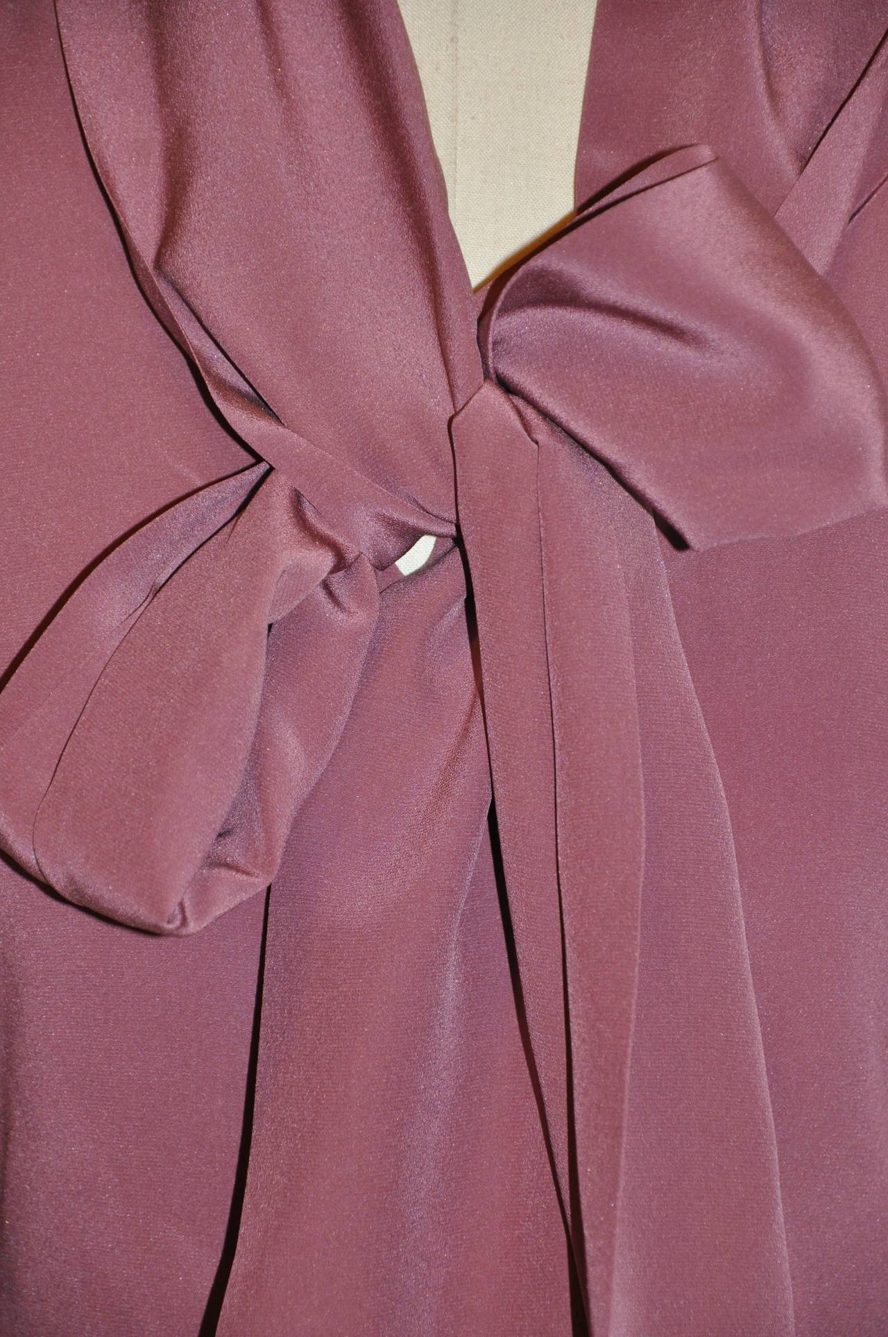 La blusa Yves Saint Laurent Rive Gauche de color malva oscuro con cuello alargado, corbata y botones delante, de crepé de chine de seda, tiene cuatro (4) botones de nácar delante, así como dos en cada puño de las mangas, que miden 5 cm de ancho. La
