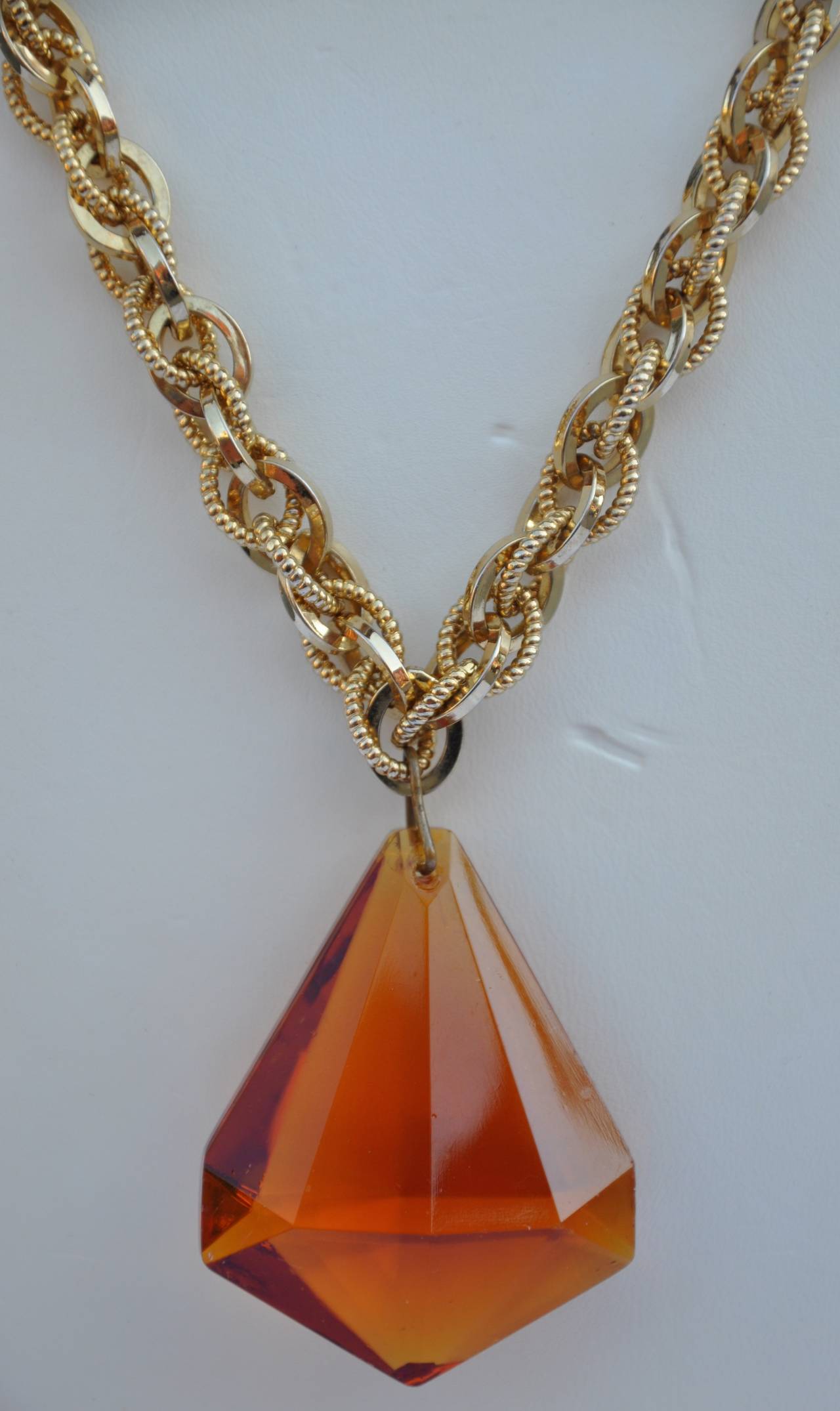 This large amber-tone lucite "Diamond" pendant measures 2" in height, 1 5/8" in length across the bottom and 6/8" in depth. The multi-textured gilded gold hardware necklace measures 18" in length.