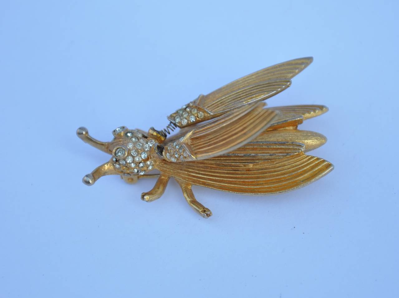            This whimsical gilded gold tone "bug" brooch has wings helped by "springs", are movable. The eyes are accented with rhinestones with detailed engraved body and wings. The total length measures 2 1/4", width is 1
