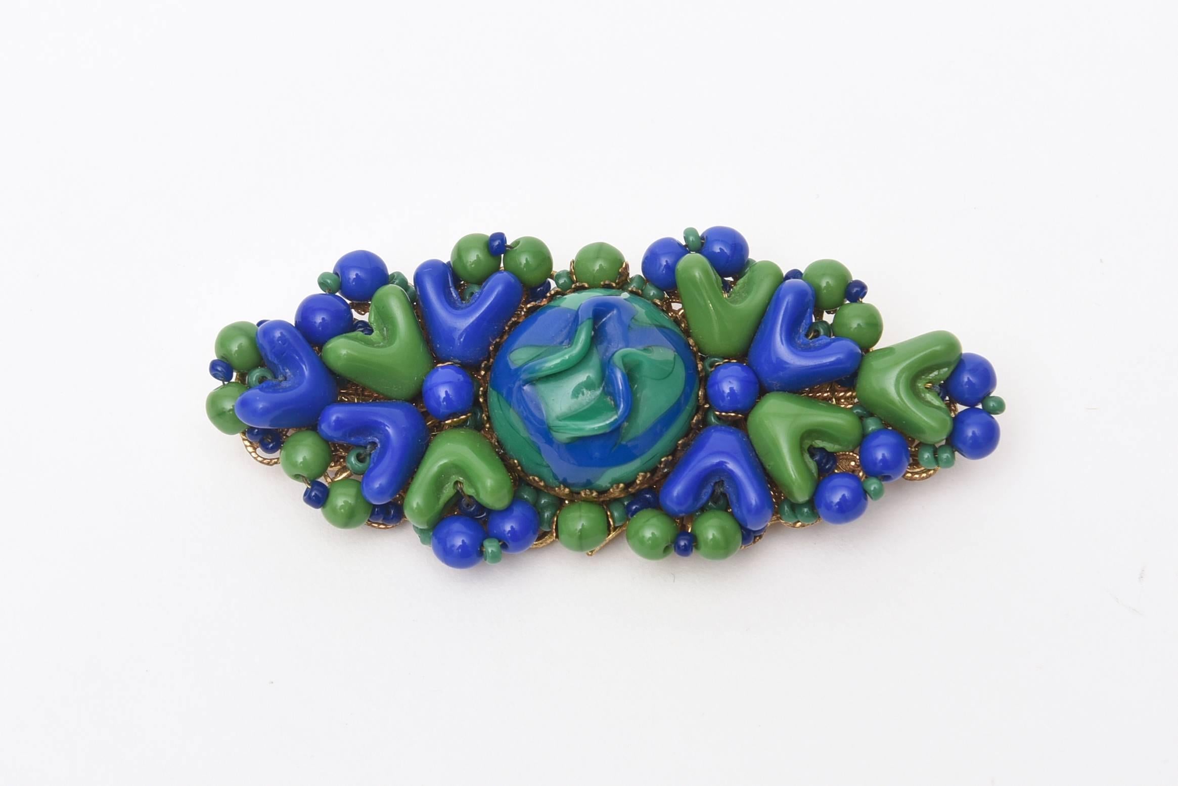 Le magnifique jeu de teintes brillantes de bleu et de vert fait de cette broche vintage signée Miriam Haskell un véritable coup de cœur.  Elle est posée sur du métal tordu. Le centre du médaillon est une combinaison des deux couleurs. Les couleurs