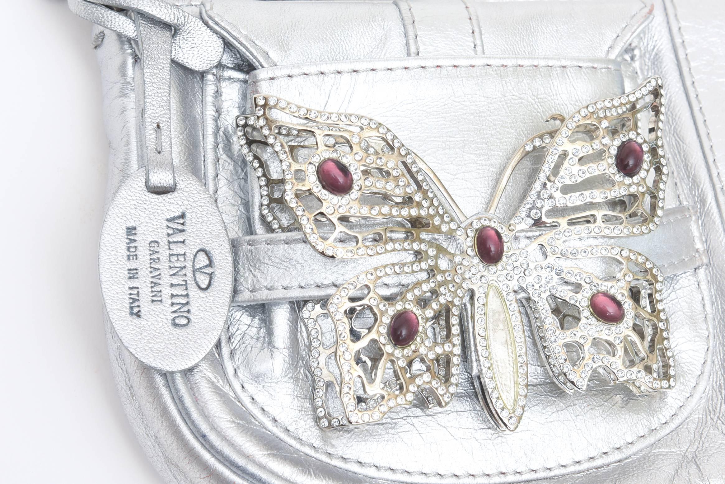 This lovely Valentino silver shoulder carrying handbag bag for day or evening is in the ever neutral silver lambskin leather with a dramatic purple and clear rhinestone butterfly for good luck.
It is from the 90's and has a front flip pocket and a