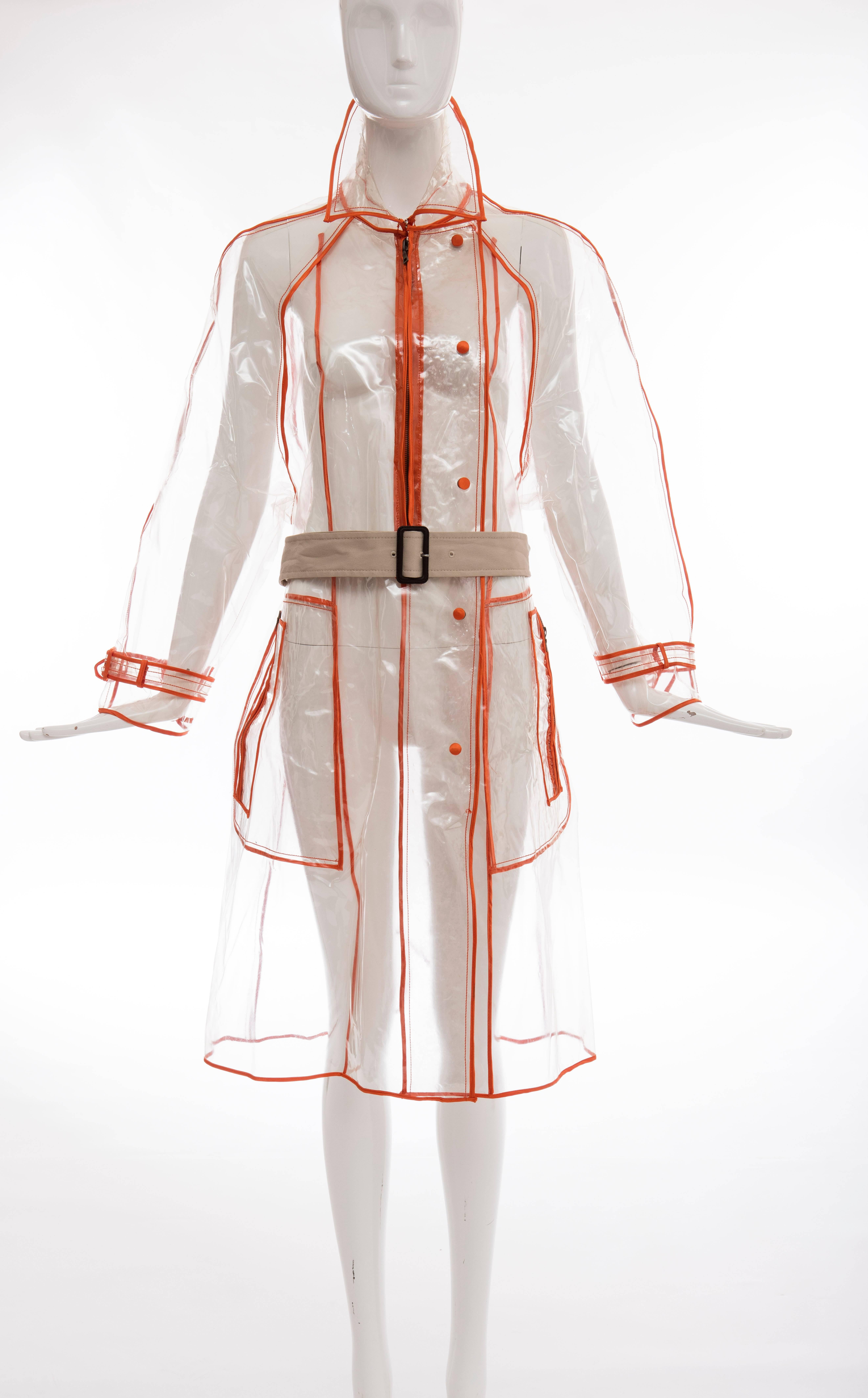 Prada, Herbst - Winter 2002-3, transparenter PVC-Regenmantel mit spitzem Kragen, langen Ärmeln, zwei seitlichen Reißverschlusstaschen, Gürtel in der Taille, kontrastierendem orangefarbenem Besatz und Druckknopf- und Reißverschlussverschlüssen vorne.