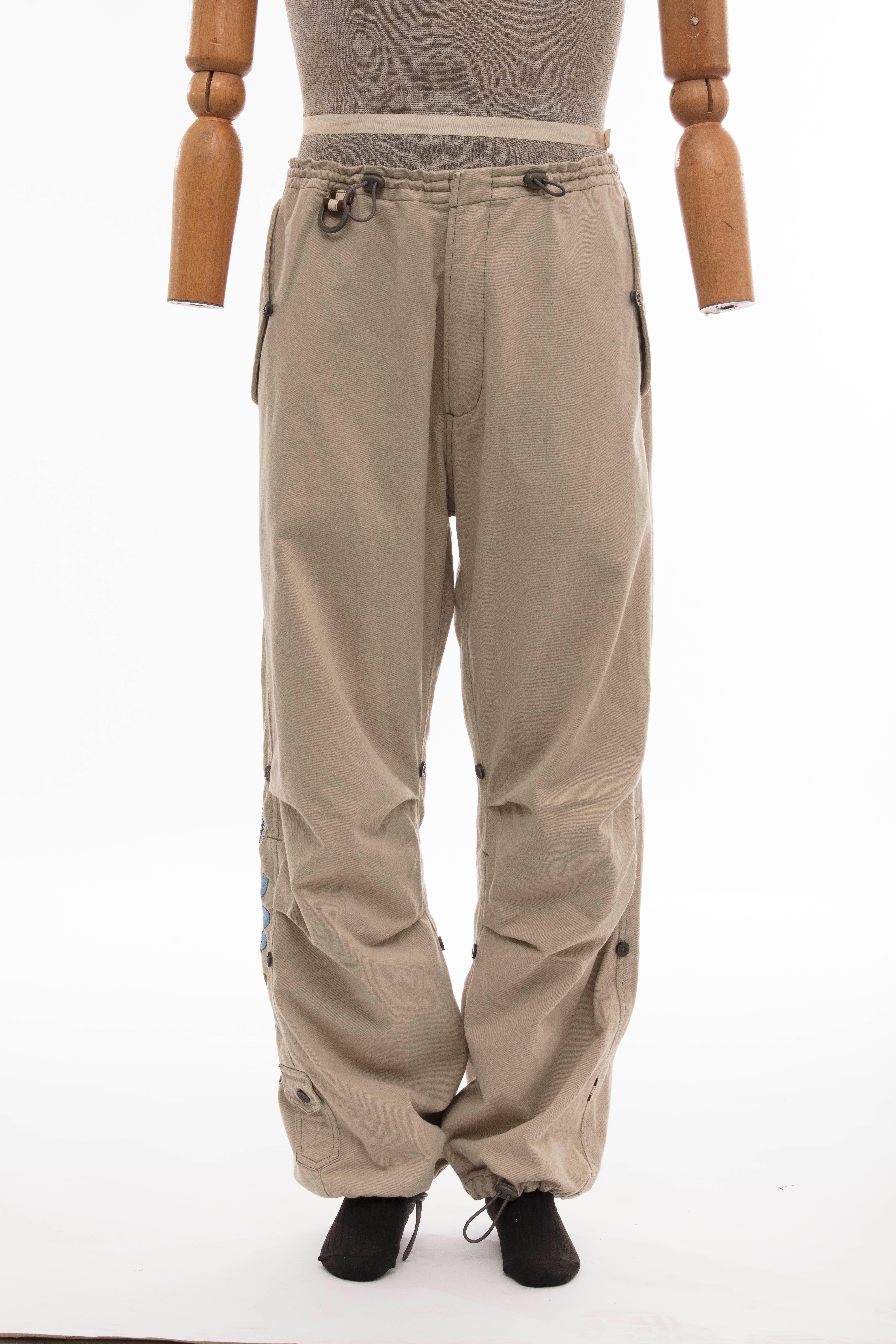  Maharishi, Circa 2004 men's khaki cotton utility pants with dual slit pockets at sides, single flap pocket at back, embroidered detail at back and drawstring pulls at waist and ankles.

Size: XL

Waist: 36, Rise 13.5, Inseam 30, Leg Opening 14

