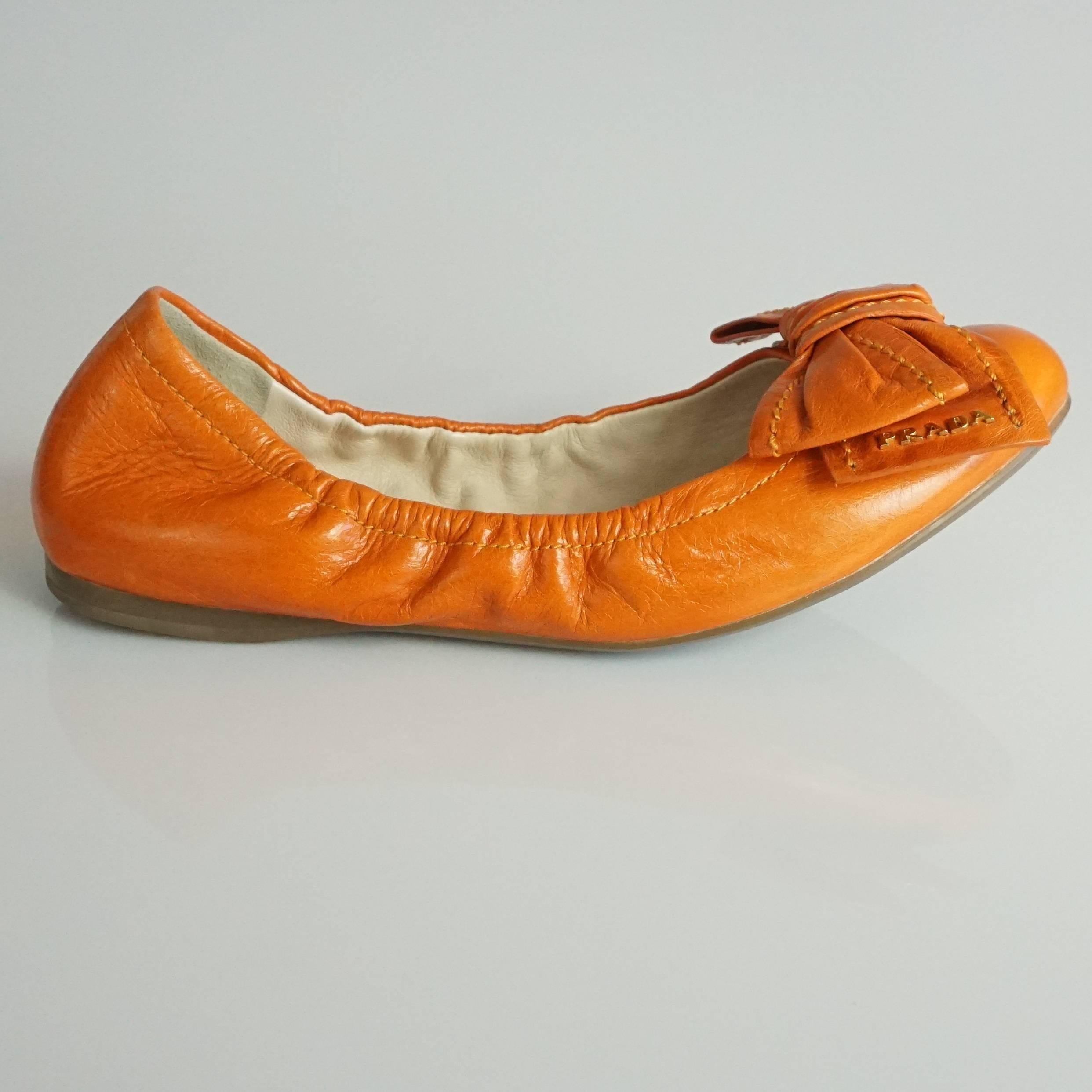 These Prada stretchy ballet flats are orange leather. They have a bow on the front and gold letters spelling out "Prada" on the bow. They are in excellent condition with some scratches / wear on the back.