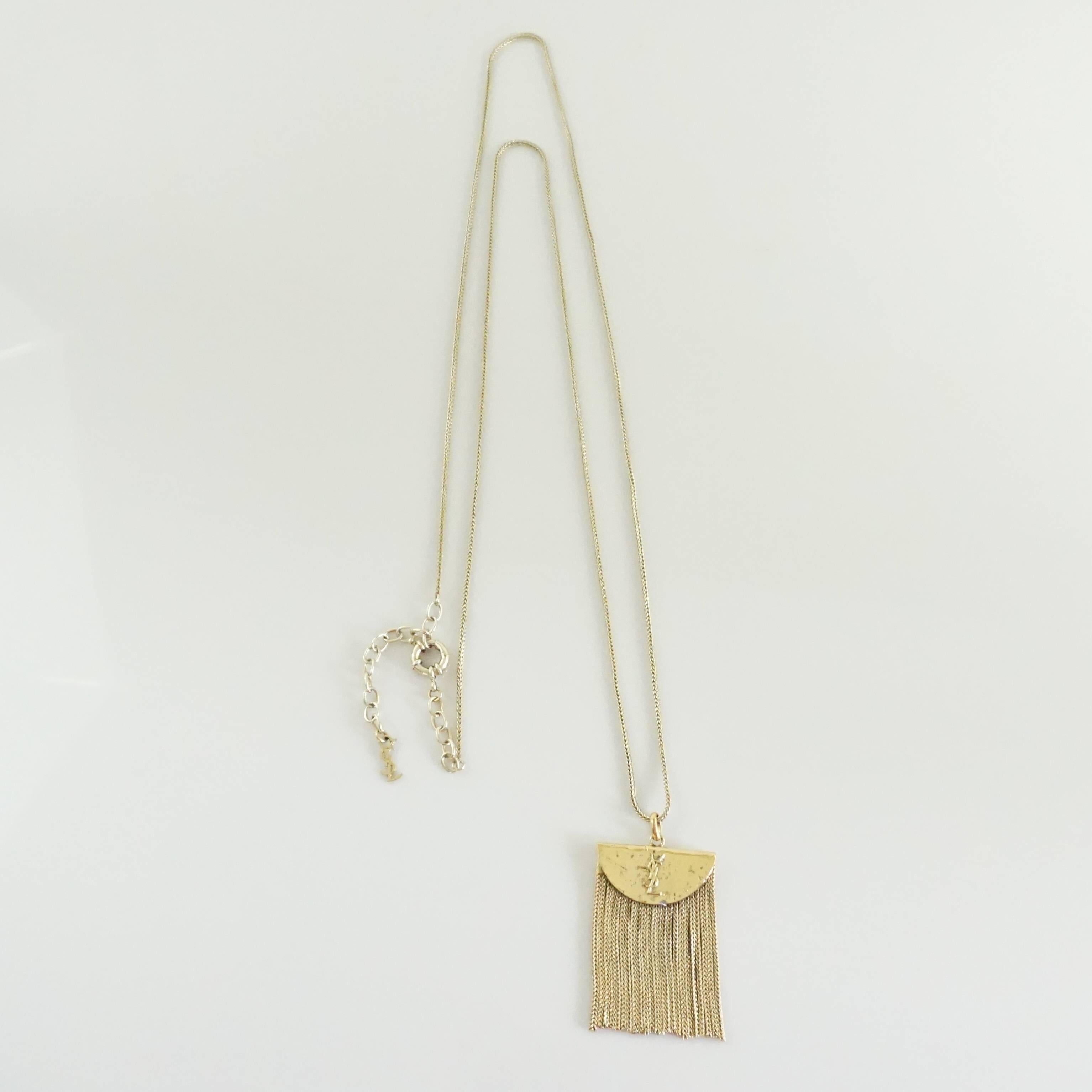 This Yves Saint Laurent long pendant necklace is a golden brass color. The pendant is a textured semi-circle with "YSL" embossed and a fringe hanging from it. This necklace is in excellent condition with some wear to the closure.
