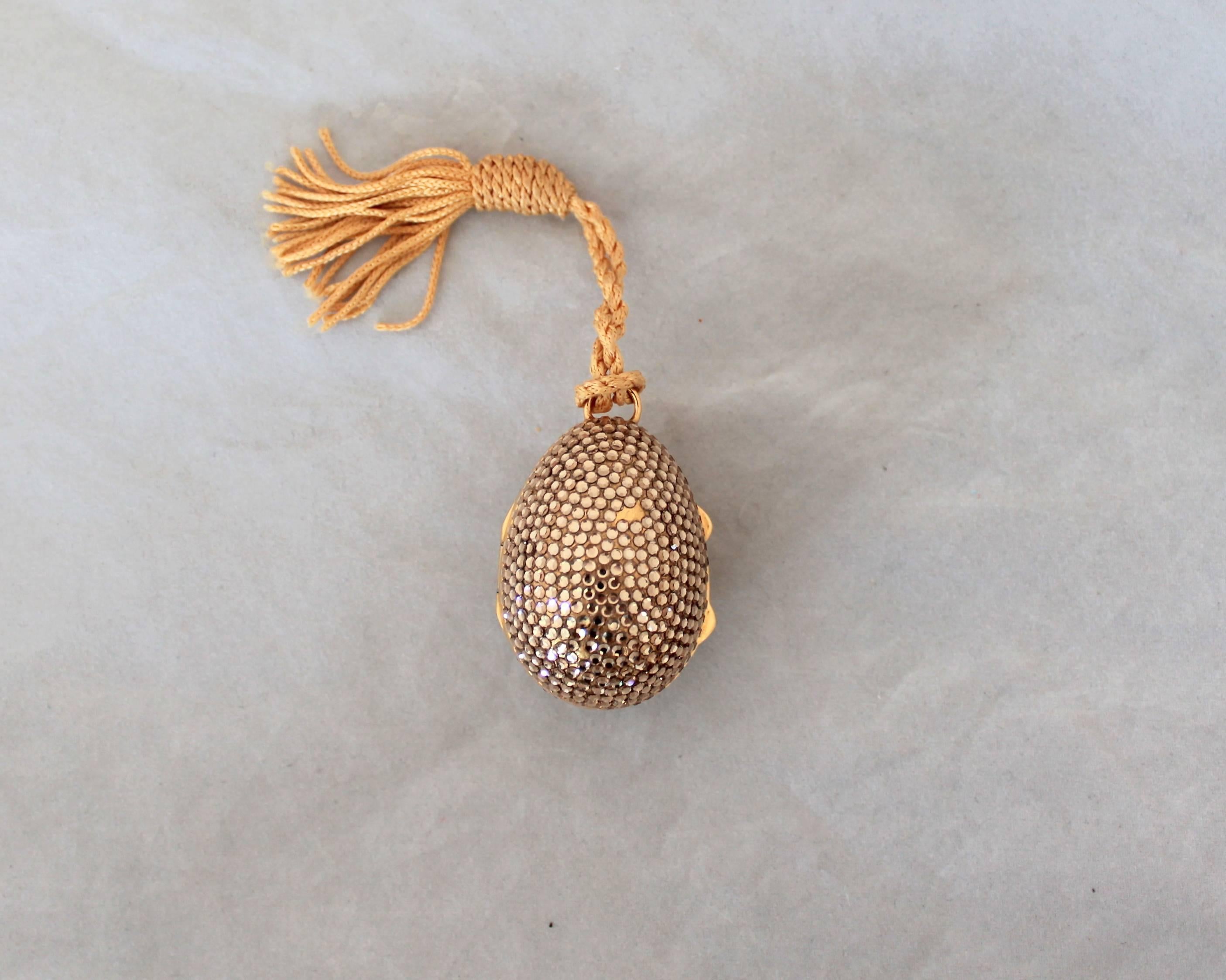 Judith Leiber Gold & Clear Rhinestone Egg Pillbox with Tassel. This piece is in fair condition with stones missing on the front and back as seen in the images. The front of the egg has a bow image.

Measurements:
Heigth- 1.75