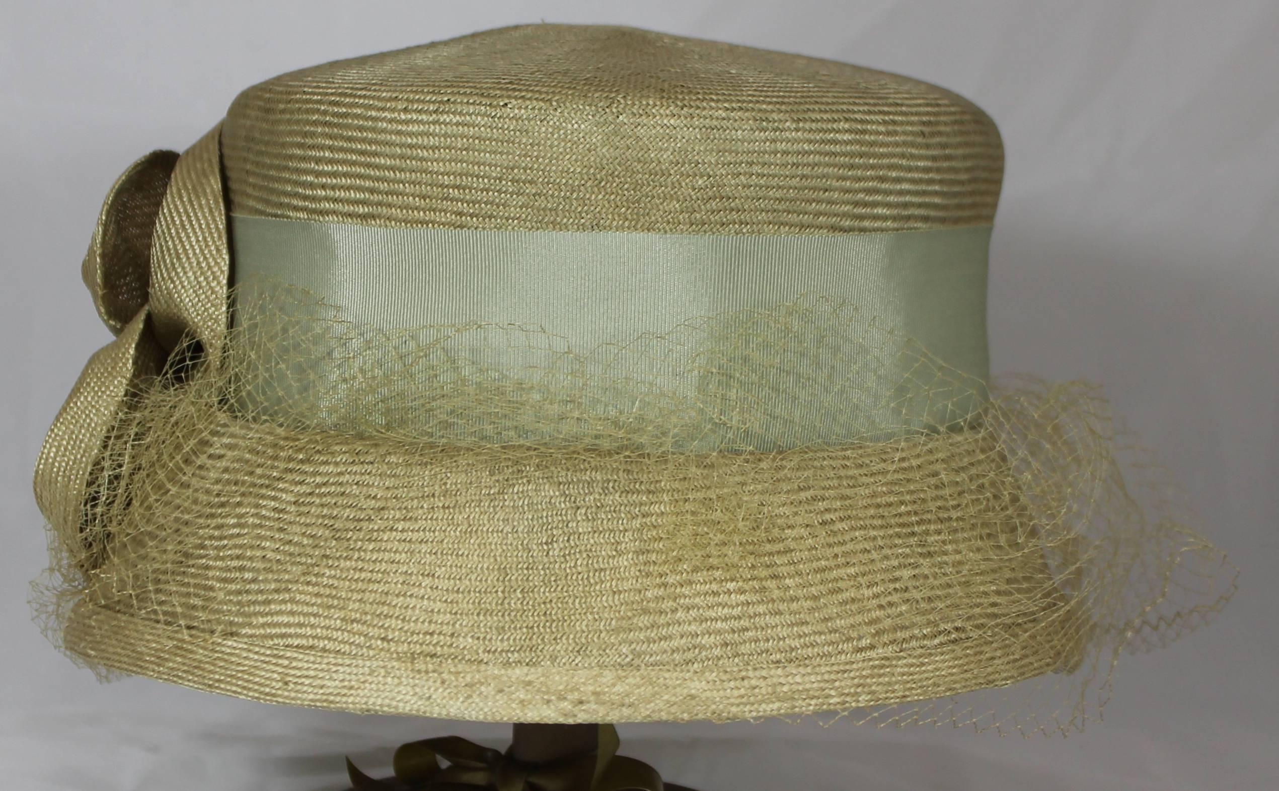 Suzanne Couture Millinery Light Olive Straw Hat with Ribbon, Flower, and Net. This hat is made of straw-like material. It is a light olive color and has a blueish green ribbon around it. There is a large light olive flower wit a button in the middle
