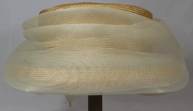 Suzanne Couture Millinery Tan Straw Hat with Synthetic Mesh Ribbon. This hat is made of a straw-like material and hat a thick synthetic mesh ribbon going around it. The ribbon comes together into a large flower bow. The hat is in very good condition
