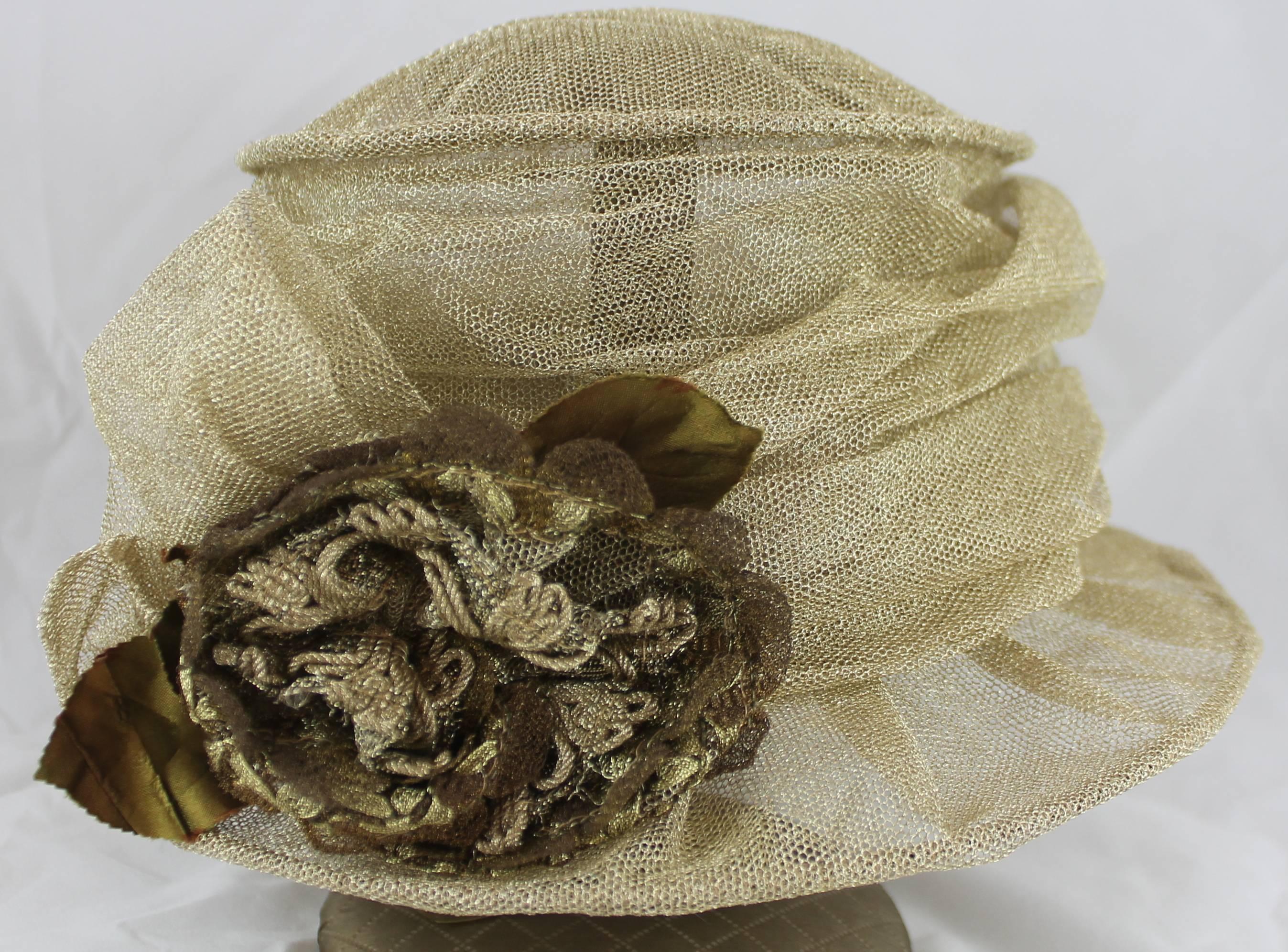 Suzanne Custom Millinery Gold Mesh Hat with Large Front Flower. This mesh hat has a structured brim. The front on the hat has a large flower and a gathering of the ribbon. This hat is in excellent condition with very light