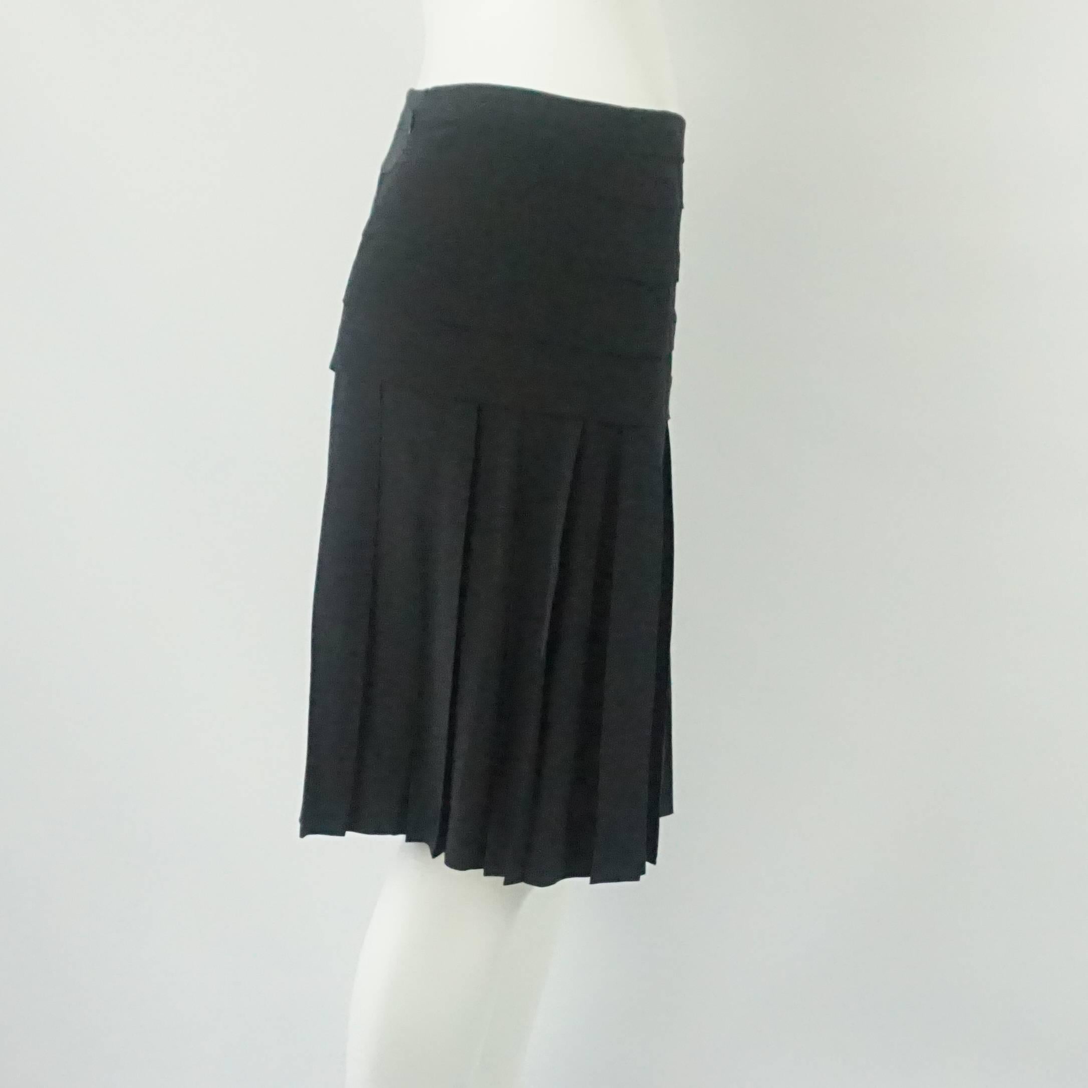 Chanel Black Silk Skirt - 38 - NWT. This Chanel skirt is black and made of silk. It is fitted around the hips and then flares out with pleating. There is a black Chanel logo detail. This skirt is in good condition and NWT with some small presses