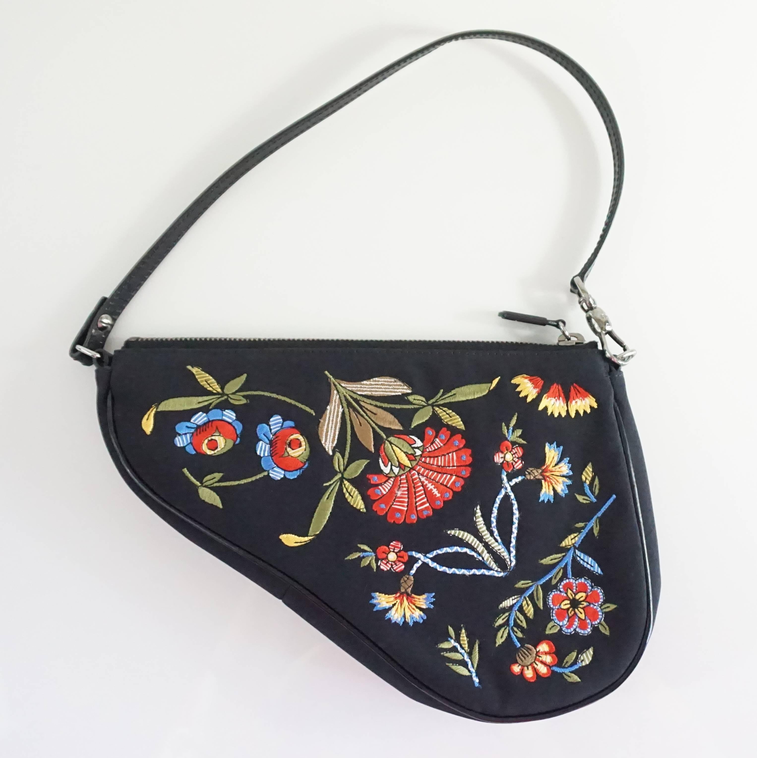 This unique Christian Dior saddle bag is black with a multicolored embroidered floral design. It has a black leather strap and a black pipe-like trim. It has a silver zipper and silver hardware. This bag is in excellent