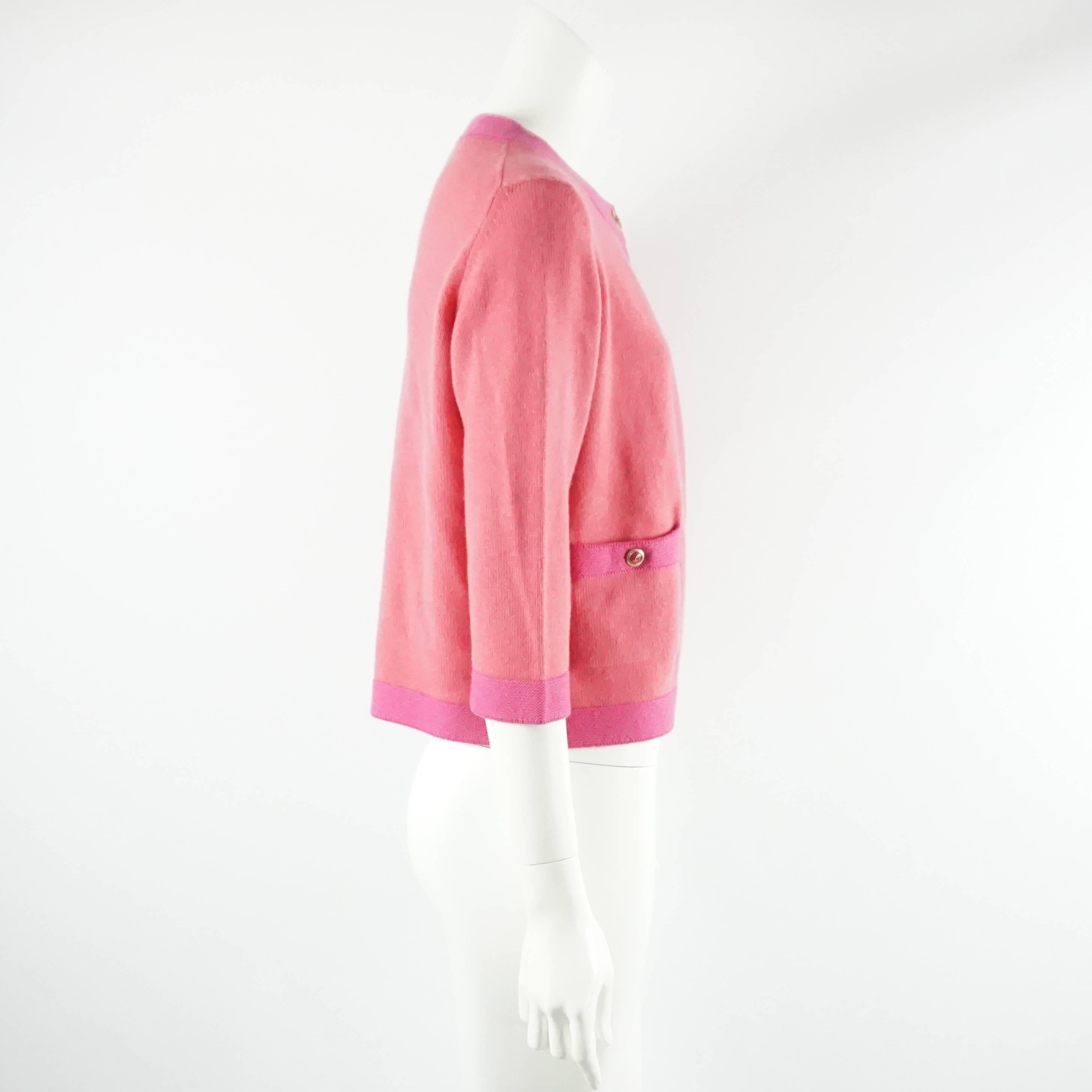 This Chanel cashmere sweater is salmon colored with a pink trim and is from the 2007P collection. There are 2 pockets on the front of the cardigan with  gold and pink Chanel logo buttons and a single top button for closure. This sweater is in