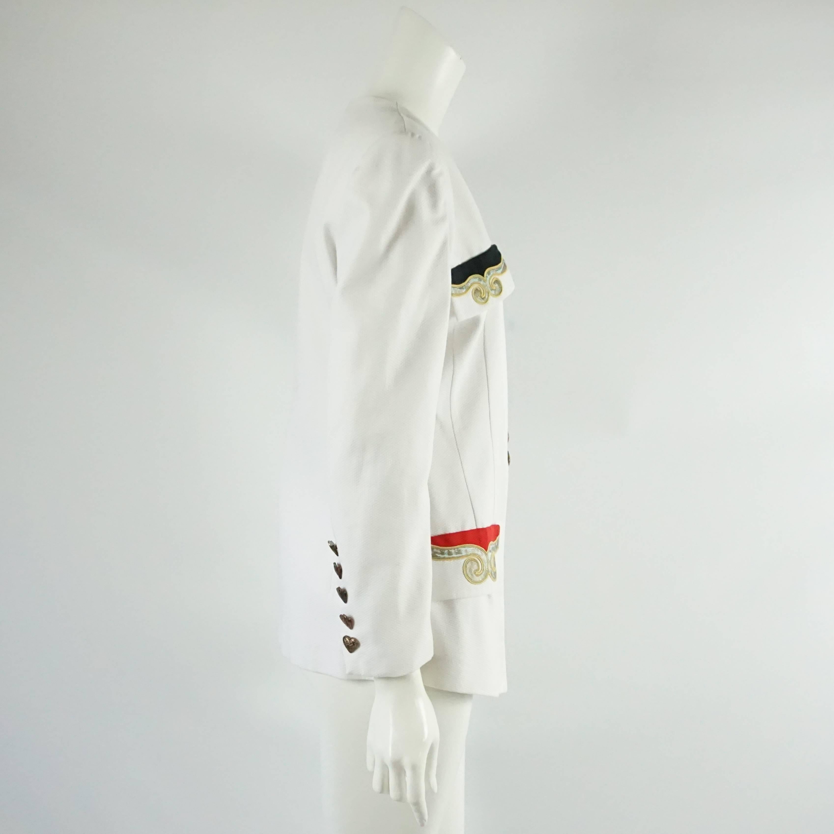 Cette veste blanche de Pierre Balmain des années 1990 comporte 4 poches sur le devant. Les poches sont brodées en rouge ou en noir avec de l'or. Cette veste est en très bon état vintage et comporte 8 boutons en bronze en forme de cœur sur le devant.