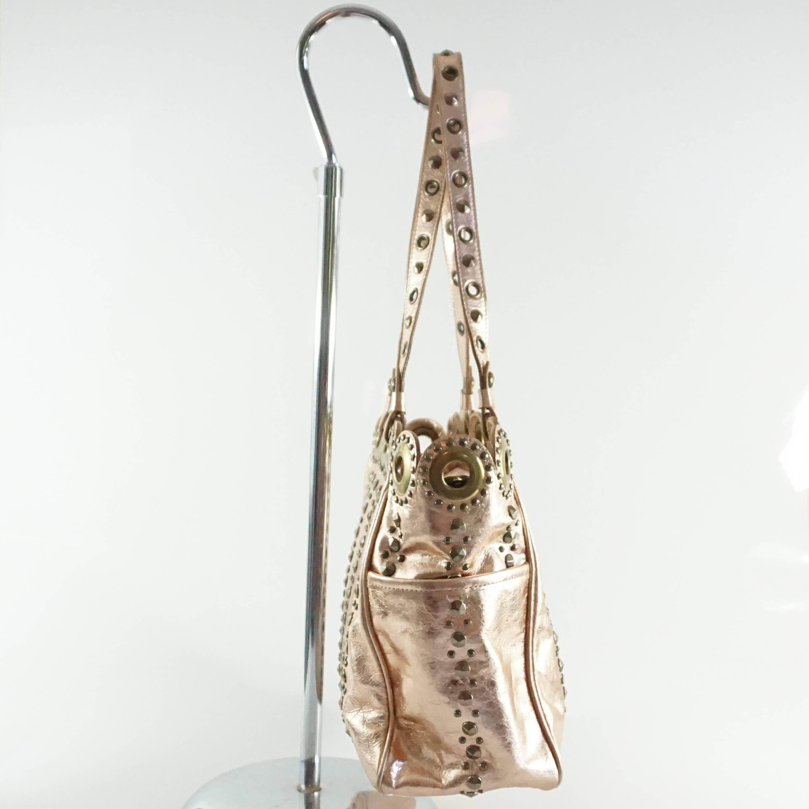 This Isabella Fiore leather shoulder bag is a metallic rose gold with dark gold studs and grommets all over. The top of the bag has a wavy shape with thin shoulder straps. The bag also has a zipper closure, 2 exterior small pockets, 1 large interior