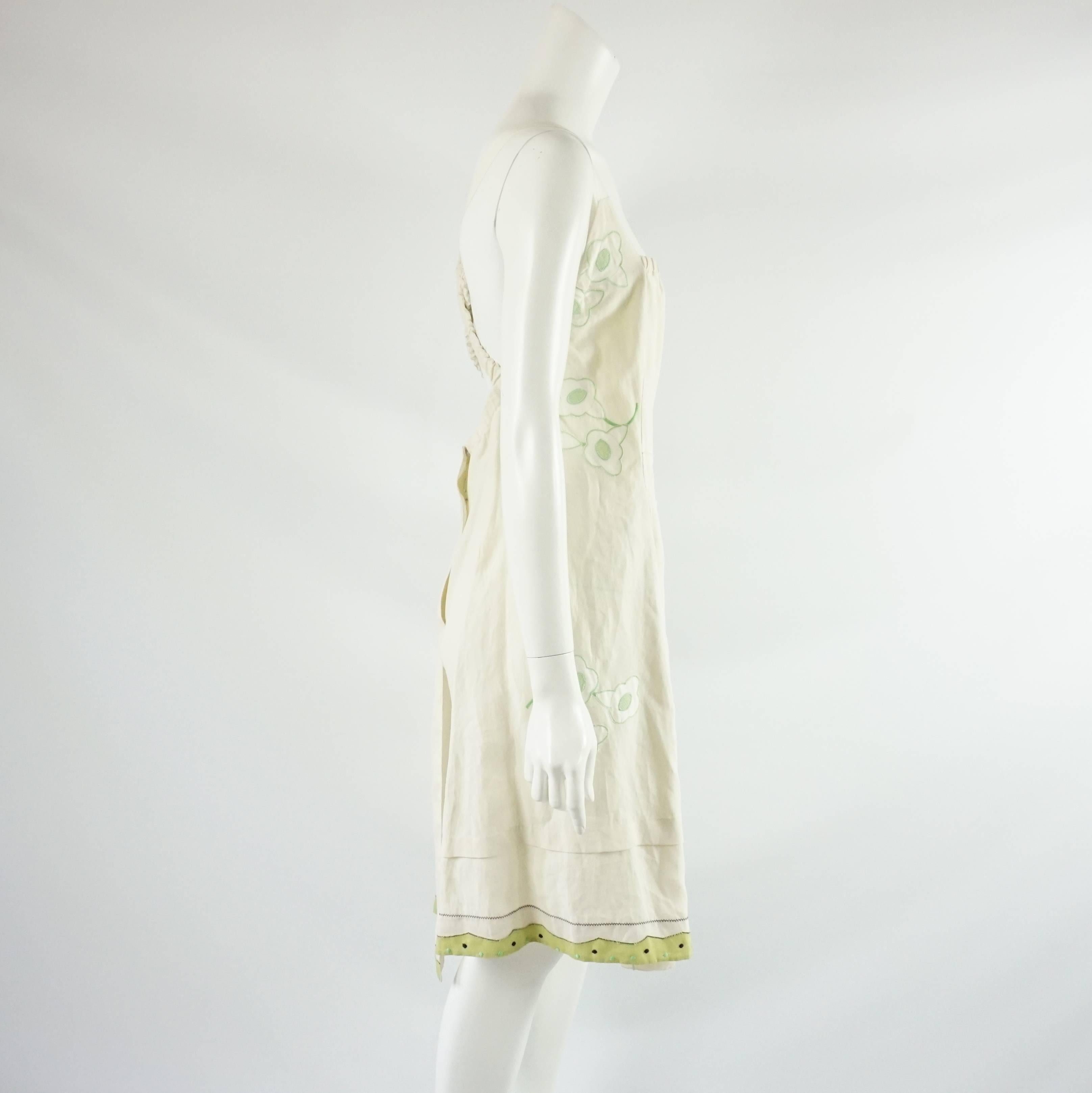 This Prada dress is ivory with green detailing. There is a floral design and dots on the trim. This dress is sleeveless with the grosgrain ribbon and elastic straps crossing each other in the back. The dress crosses over in the back with snap