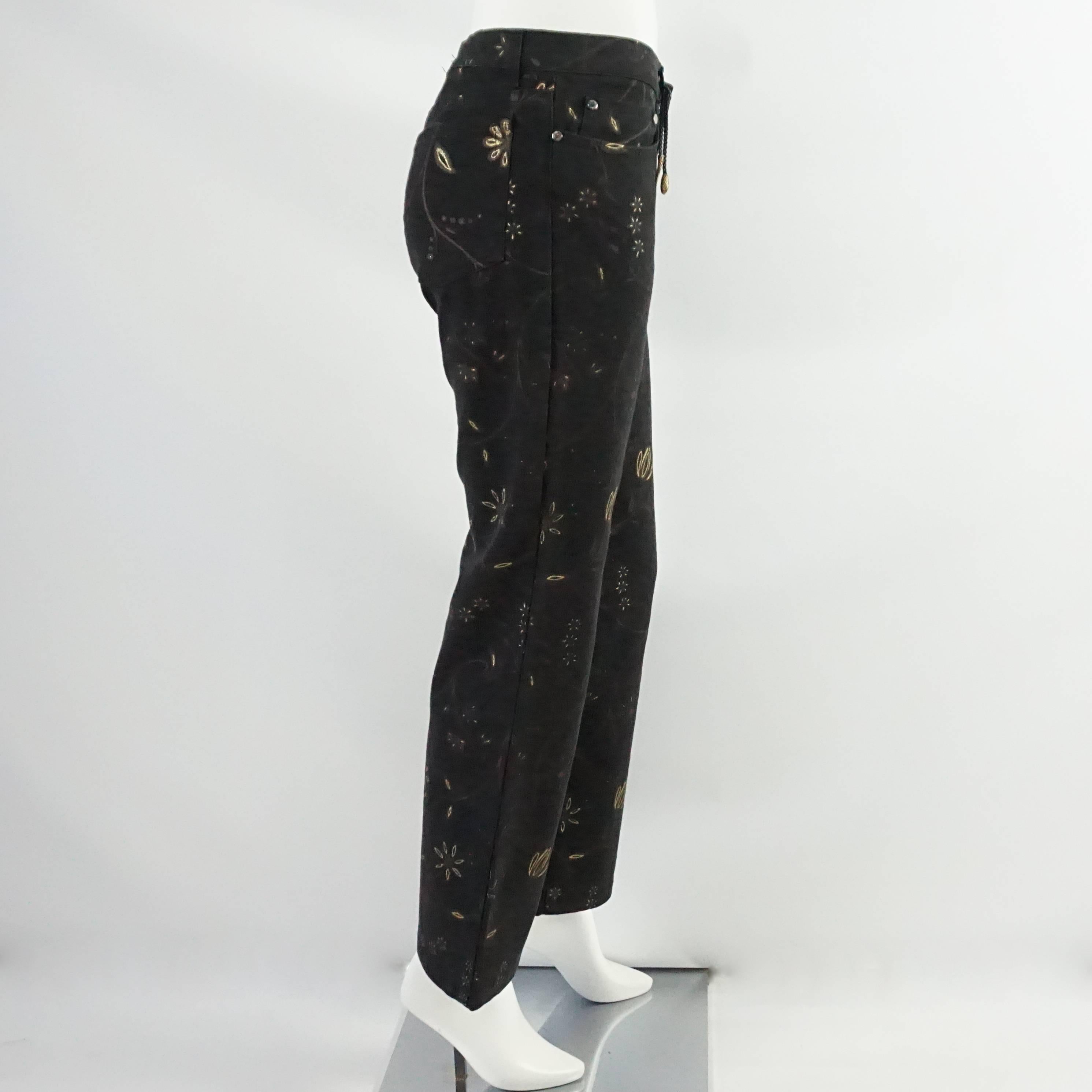 These Mid Waisted 1990's Roberto Cavalli jeans are black and have a gold metallic floral print. The hardware is silver and there is a black braided detail with 2 bronze charms (head and tail of a snake) hanging from one of the belt loops. The button