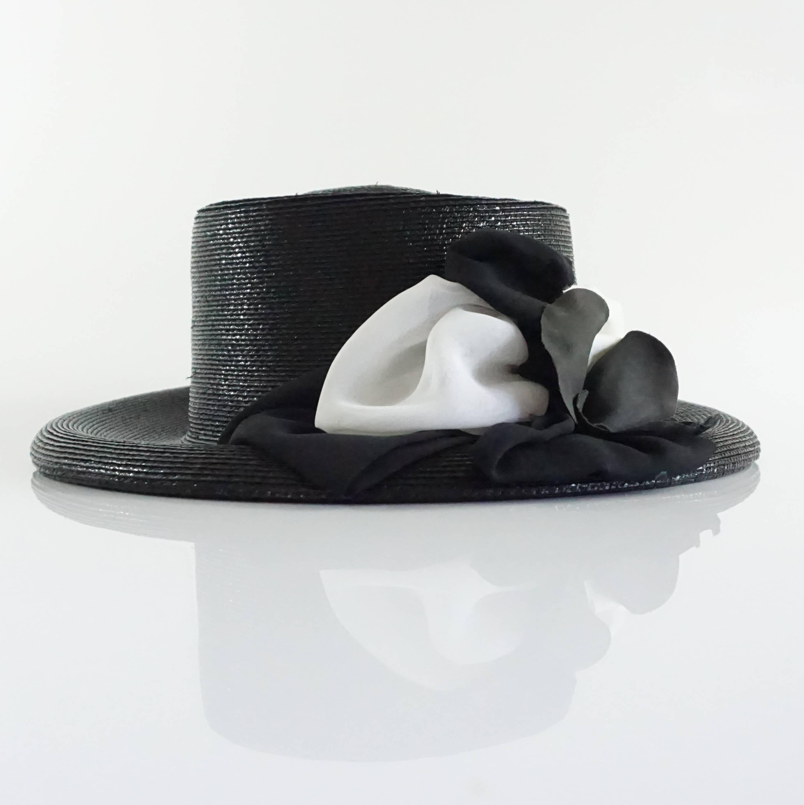 This Suzanne Couture Millinery hat is a black and glossy straw hat. The interior of the brim is white. On the outside, there is a black and white ribbon detailing along with a black and white silk flower. This hat is in excellent