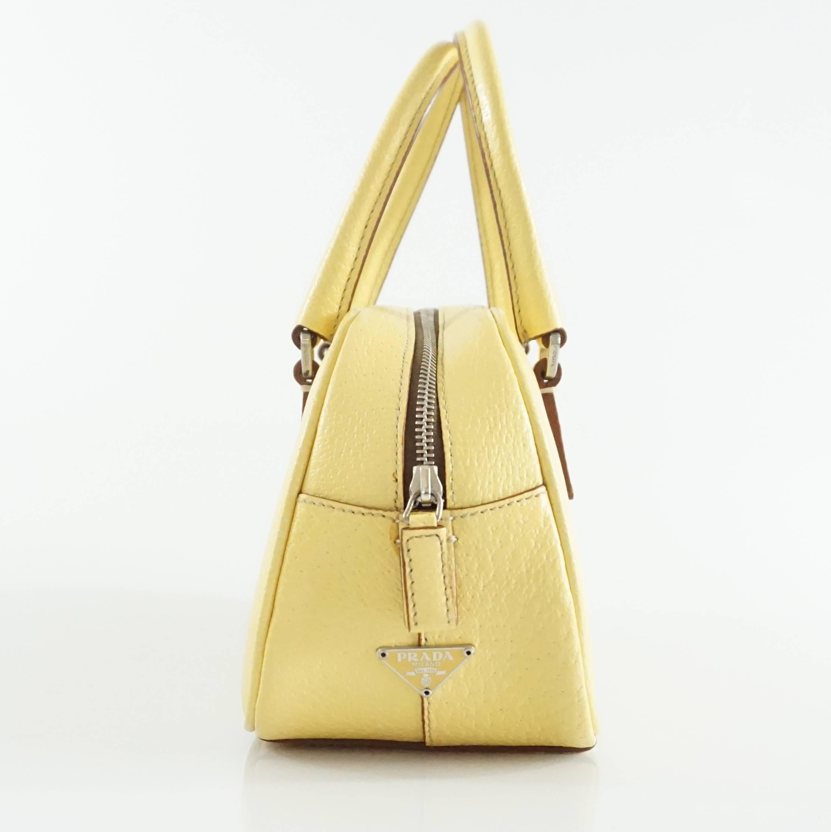 This Prada yellow top handle bag is a mini size and is leather. It has a zipper, two handle, an interior zipper pocket, and silver hardware. This bag is in fair condition with overall staining on the leather as seen in the last images.