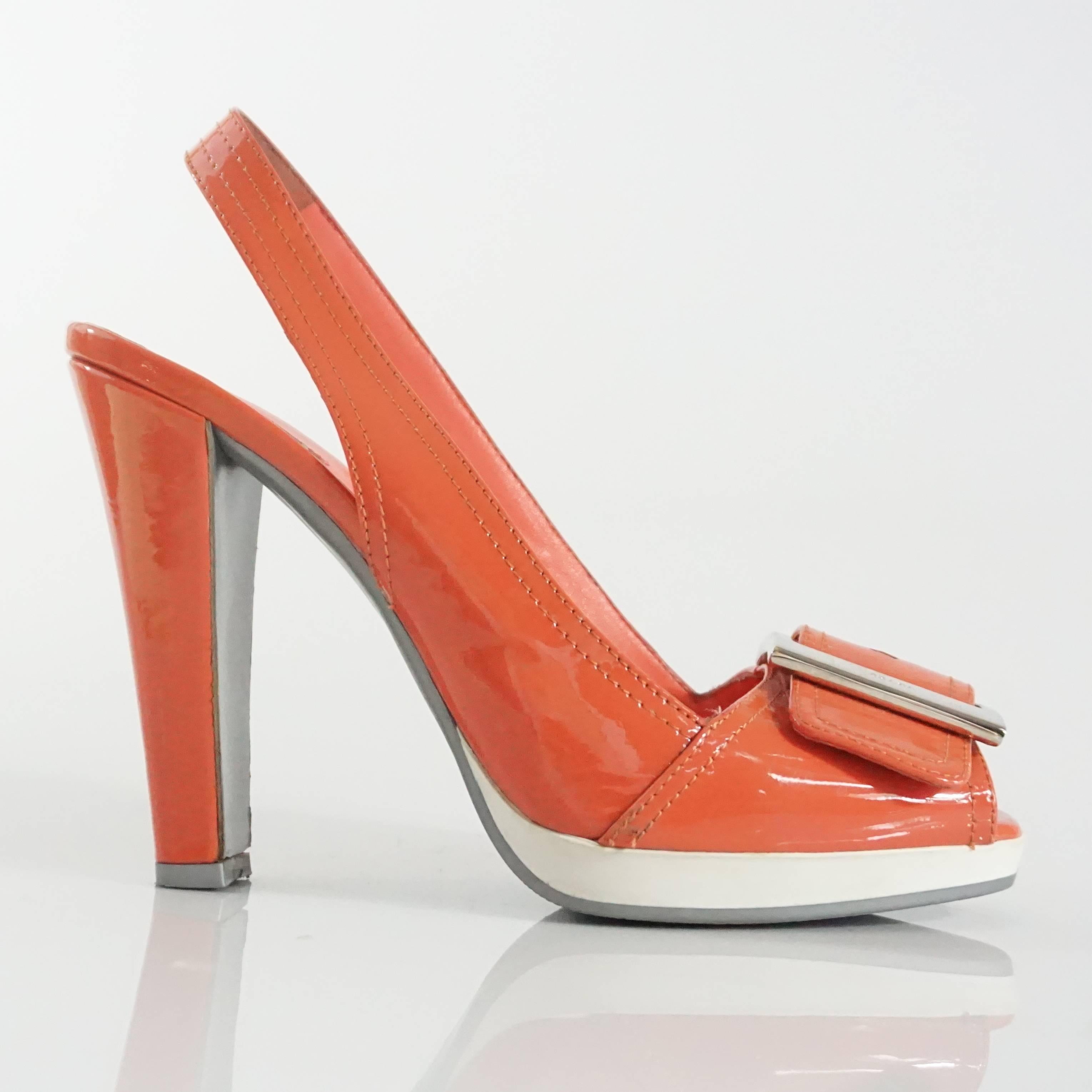 These Prada slingbacks are coral patent leather and feature a silver buckle with "Prada" engraved. There is a white platform. These heels are in very good condition with minor wear and markings on the white platform, heel, and patent
