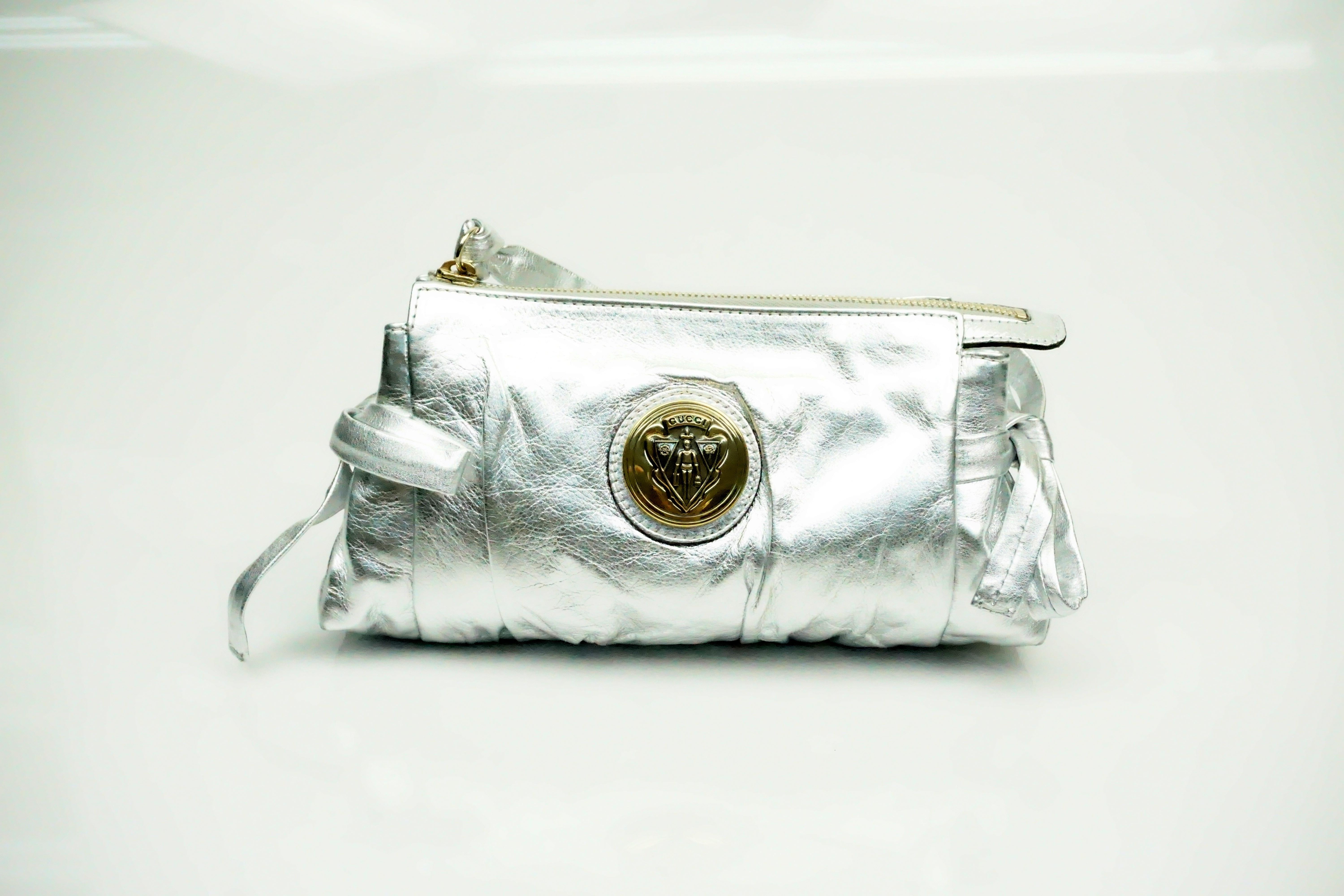 Gucci Silver Metallic Leather Clutch  This beautifulclutch is in excellent condition. The bag has two bows on the sides of the clutch. The zipper is a gold tone and has a wrist strap attached to it. There is also a gold Gucci logo on the center