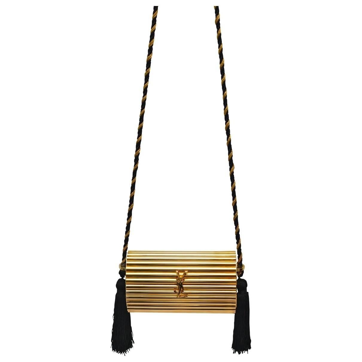 YVES SAINT LAURENT rare and collectable vintage gold tone ribbed minaudiere featuring :

- Hard box cylindric body.
- Large YSL logo at the front.
- Black and gold braided shoulder strap.
- Long black silk tassels.
- Gold canvas
