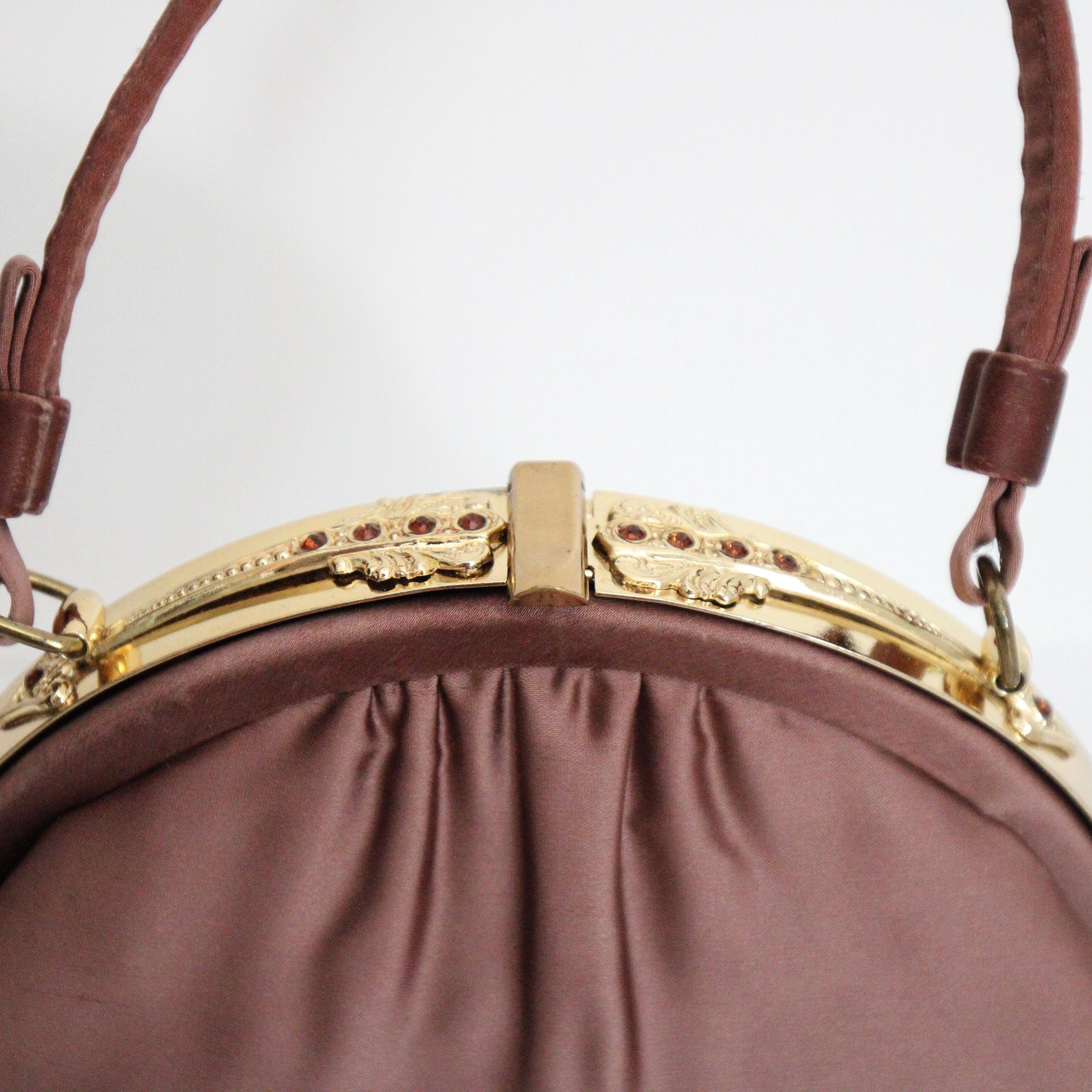 Diese wunderschöne mauvefarbene Satinhandtasche aus den 1950er Jahren, verziert mit einem verzierten Rahmen, ist das perfekte Stück, um Ihre Abendgarderobe zu vervollständigen 

Der abgerundete, goldfarbene Metallrahmen ist mit zwei Pfeilmotiven