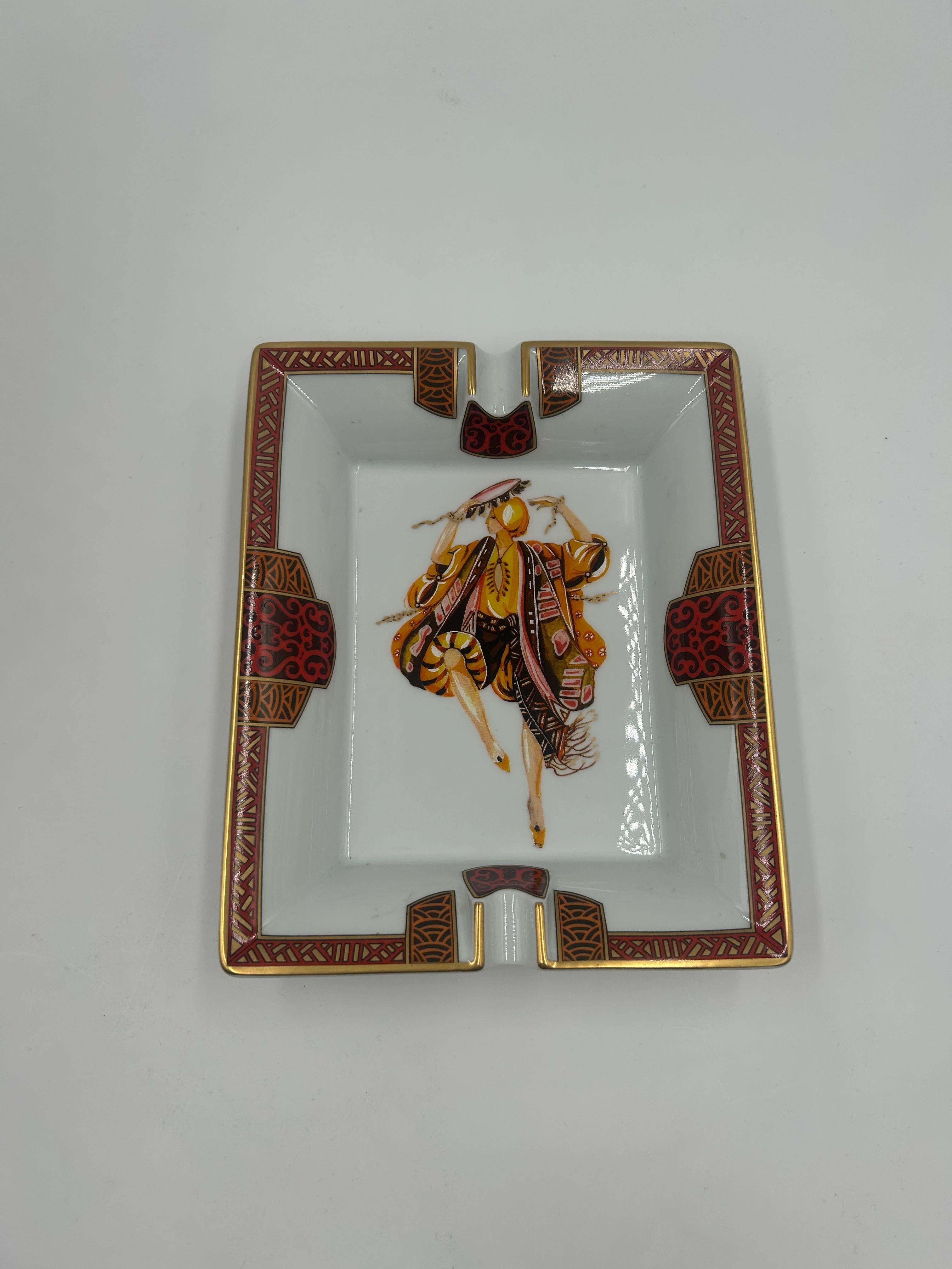 This Hermès “Dancer” Change Tray/Ashtray is a refined porcelain accessory adorned with a theatrical dancer motif in vivid, rich hues, framed by vibrant borders and polished gold accents. Crafted in France and stamped “Hermès – Paris,” the tray rests
