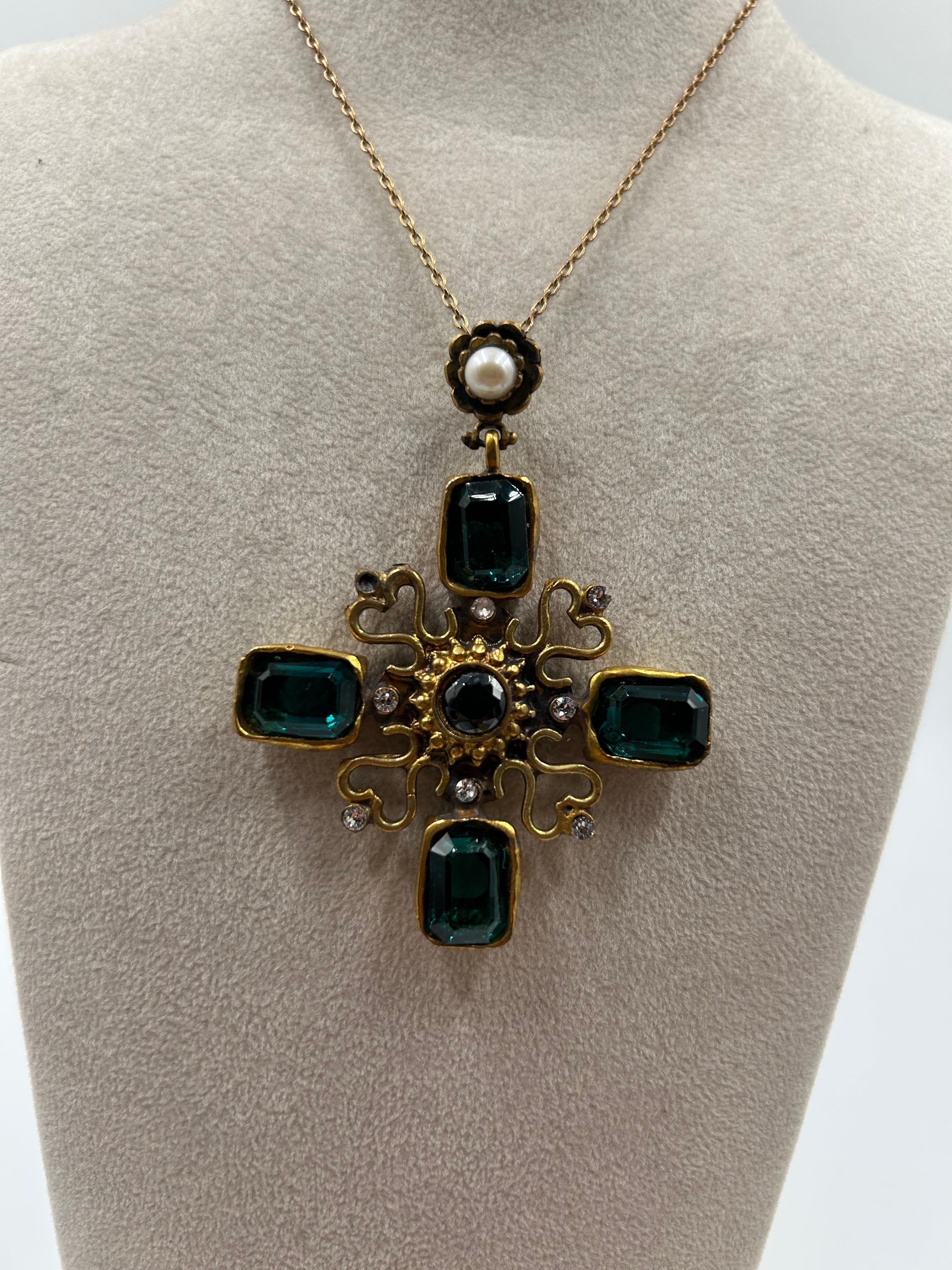 Questo ciondolo vintage in ottone dorato è caratterizzato da un audace disegno a croce ornato da pietre verdi e perle brillanti. I dettagli intricati e le texture stratificate evocano un'estetica senza tempo di ispirazione barocca, mentre il