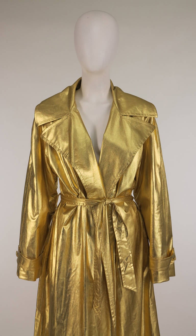 1980s Raincheetahs liquid gold trench coat...Gold wrap front coat has deep wide lapels, on seam side pockets, self wrap belt and loops...Long raglan sleeves with band and button detail at each cuff...Lightweight synthetic fabric...The coat is fully