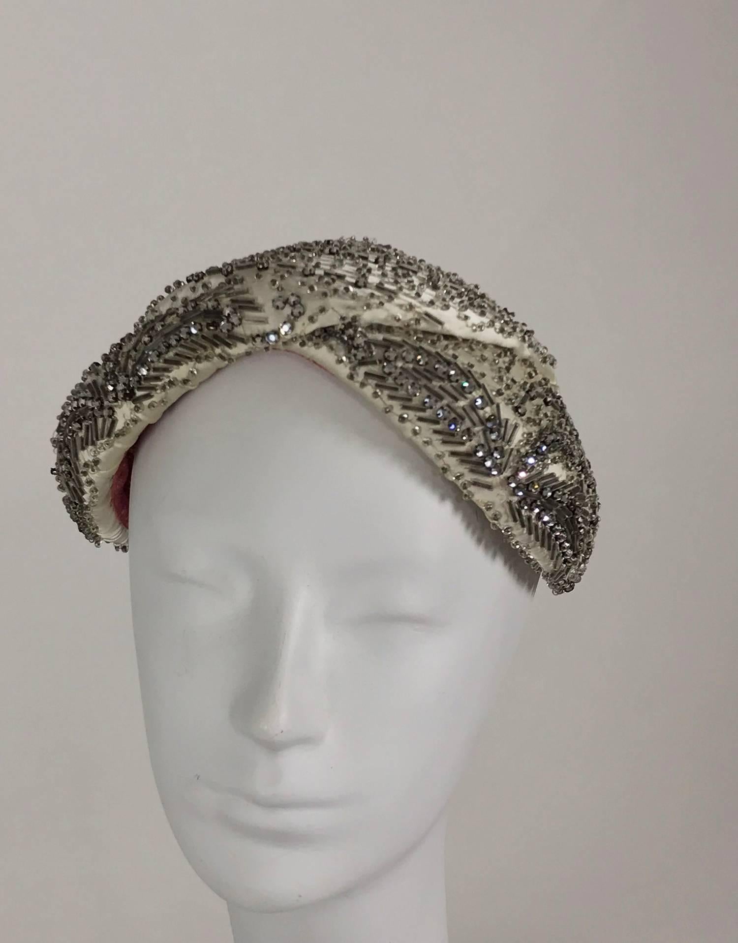 Designed by Lora rhinestone and beaded cocktail hat from the 1950s. Classic 50s band style hat of off white, with a touch of silver, silk heavily beaded in crystal and silver and set with rhinestones. The front of the hat has foliate sprays of