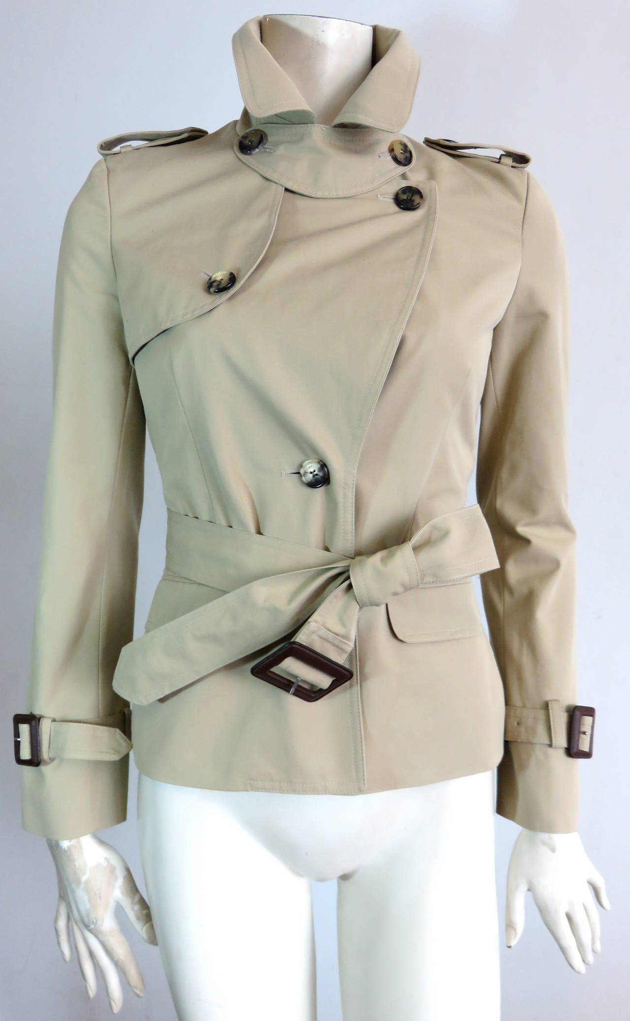 Never worn, CHRISTIAN DIOR by John Galliano cropped, cotton trench jacket.

Beige, compact woven, cotton shell fabric with silk, body lining.

Adjustable, dark brown, leather belt buckle hardwares at cuffs, and waist belt.

Logo engraved, horn